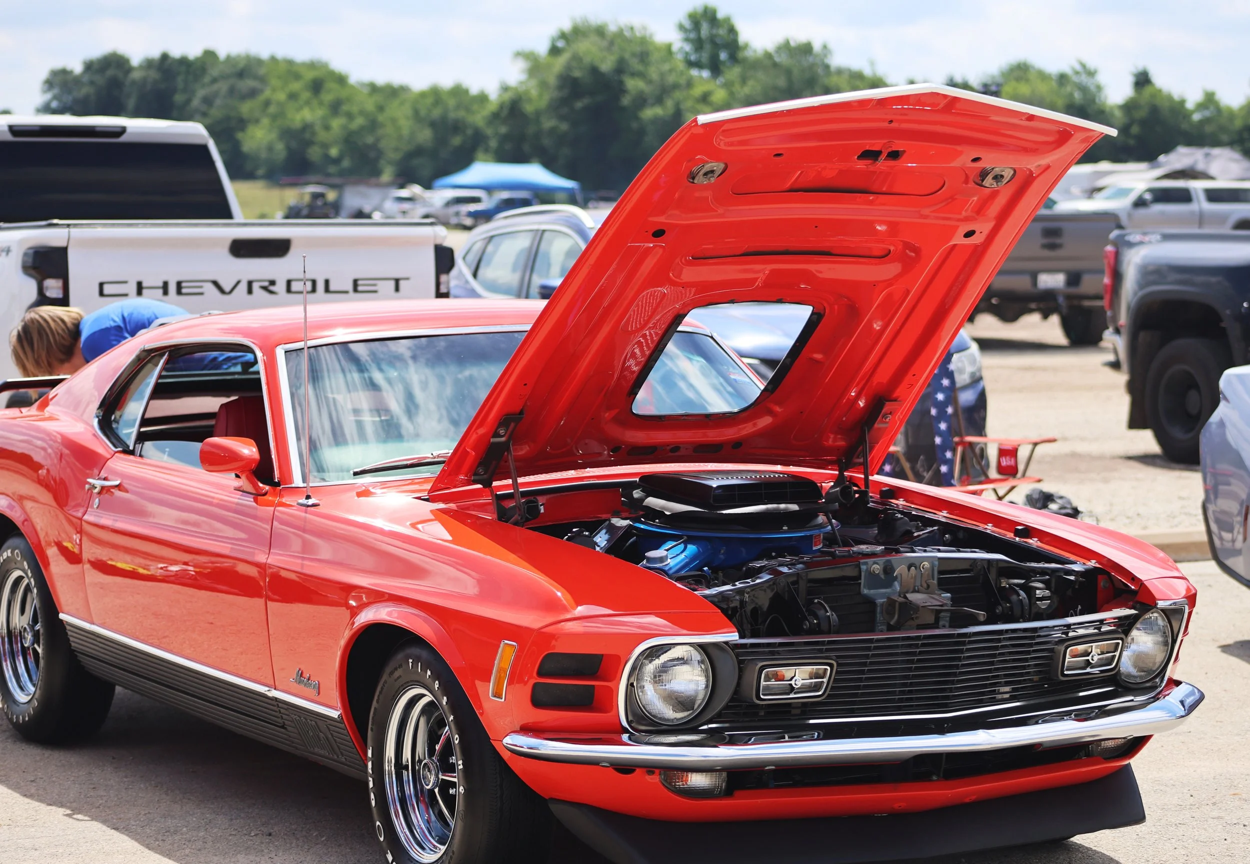 A red vintage Chevrolet muscle car with its hood open, displayed at an outdoor car show, with cars and tents in the background.