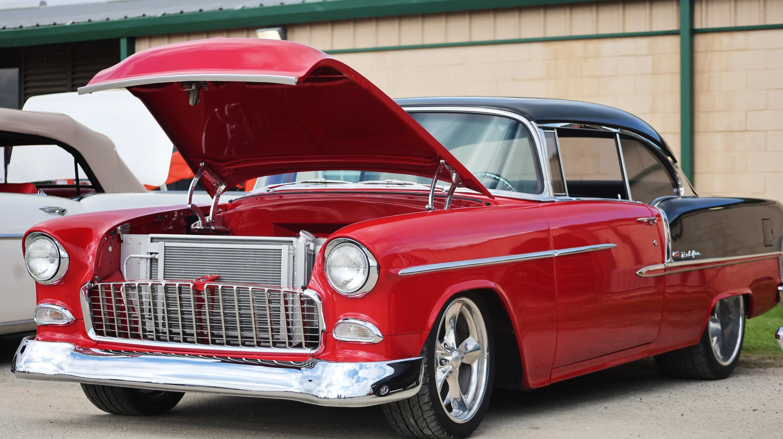 A classic red car with its hood open, showcasing the engine, parked at an outdoor car show next to other vintage vehicles.