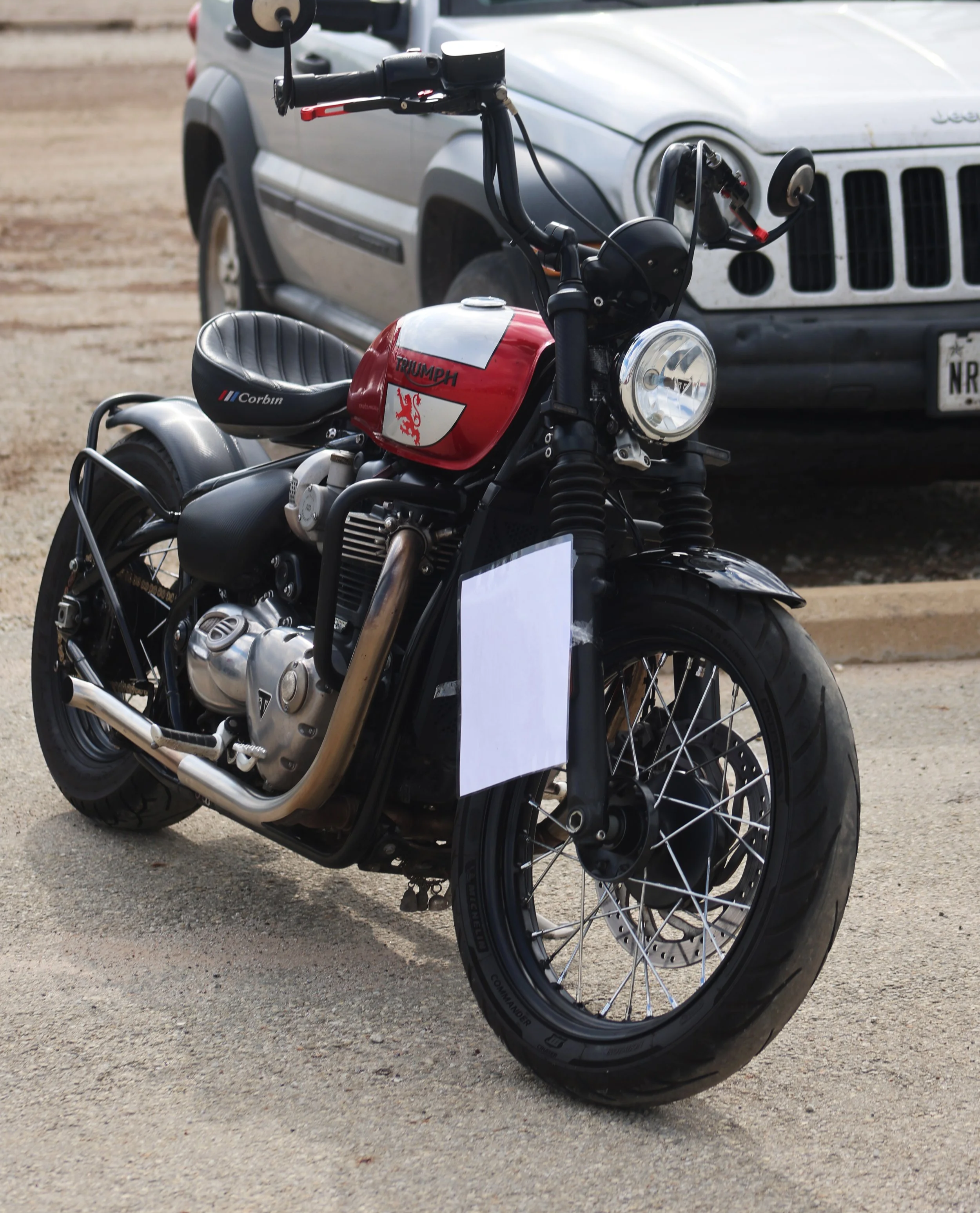 A Triumph motorcycle parked on a dirt surface in front of a gray Jeep vehicle.