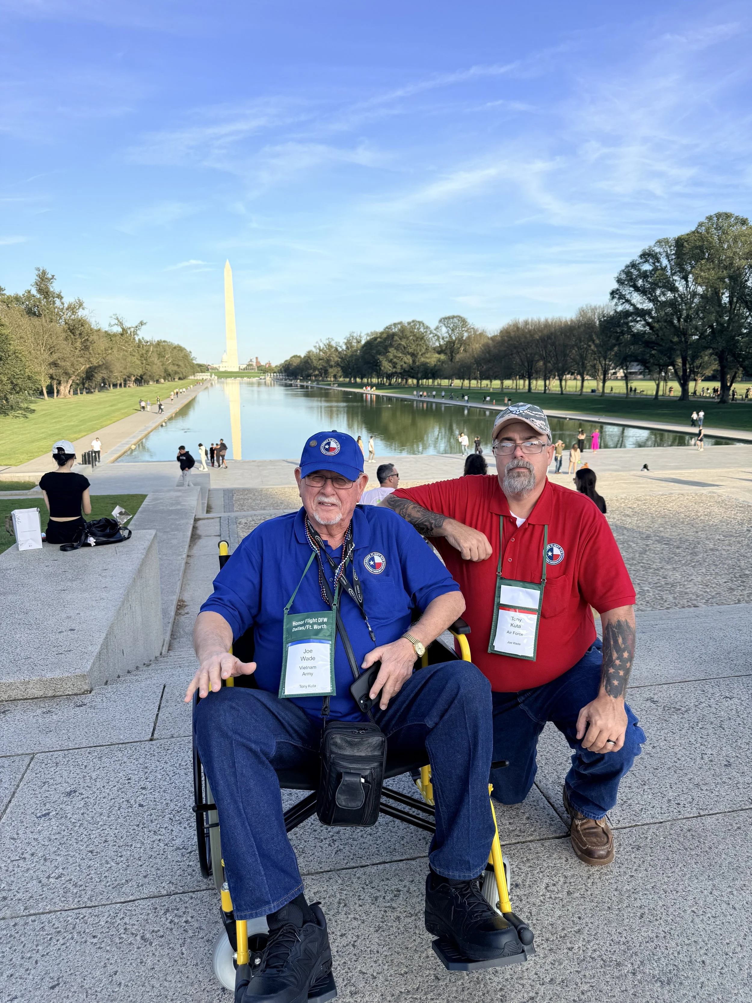Two men are sitting outdoors in front of the Lincoln Memorial Reflecting Pool, with the Washington Monument visible in the background. They appear to be at a memorial event or gathering, wearing badges and casual clothing.