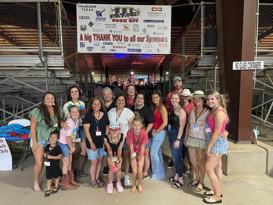 A group of 16 people, including children and adults, standing together at an outdoor event under a metal pavilion with stairs and a banner above them. They are smiling, some wearing name tags, with a stage visible in the background. The scene appears lively and festive.