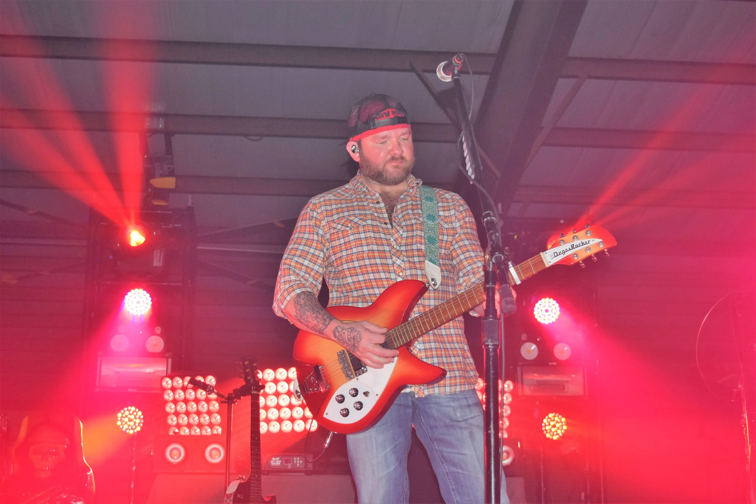 A man playing an orange electric guitar on stage during a concert, with colorful stage lights and equipment in the background.