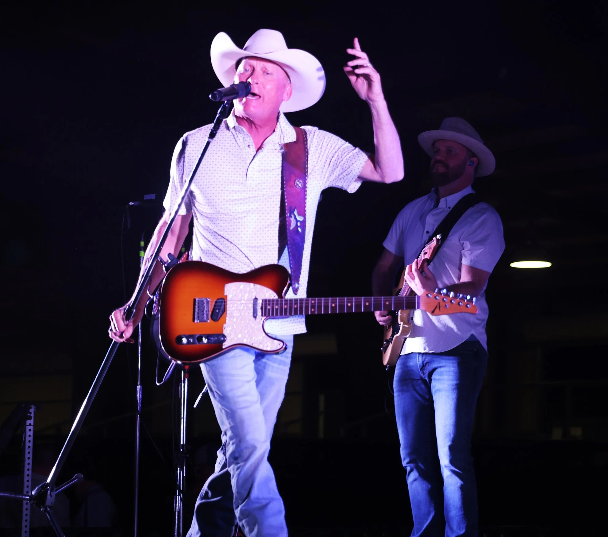 A man wearing a cowboy hat and patterned shirt singing into a microphone and playing an acoustic guitar during a musical performance, with another musician playing an electric guitar in the background.