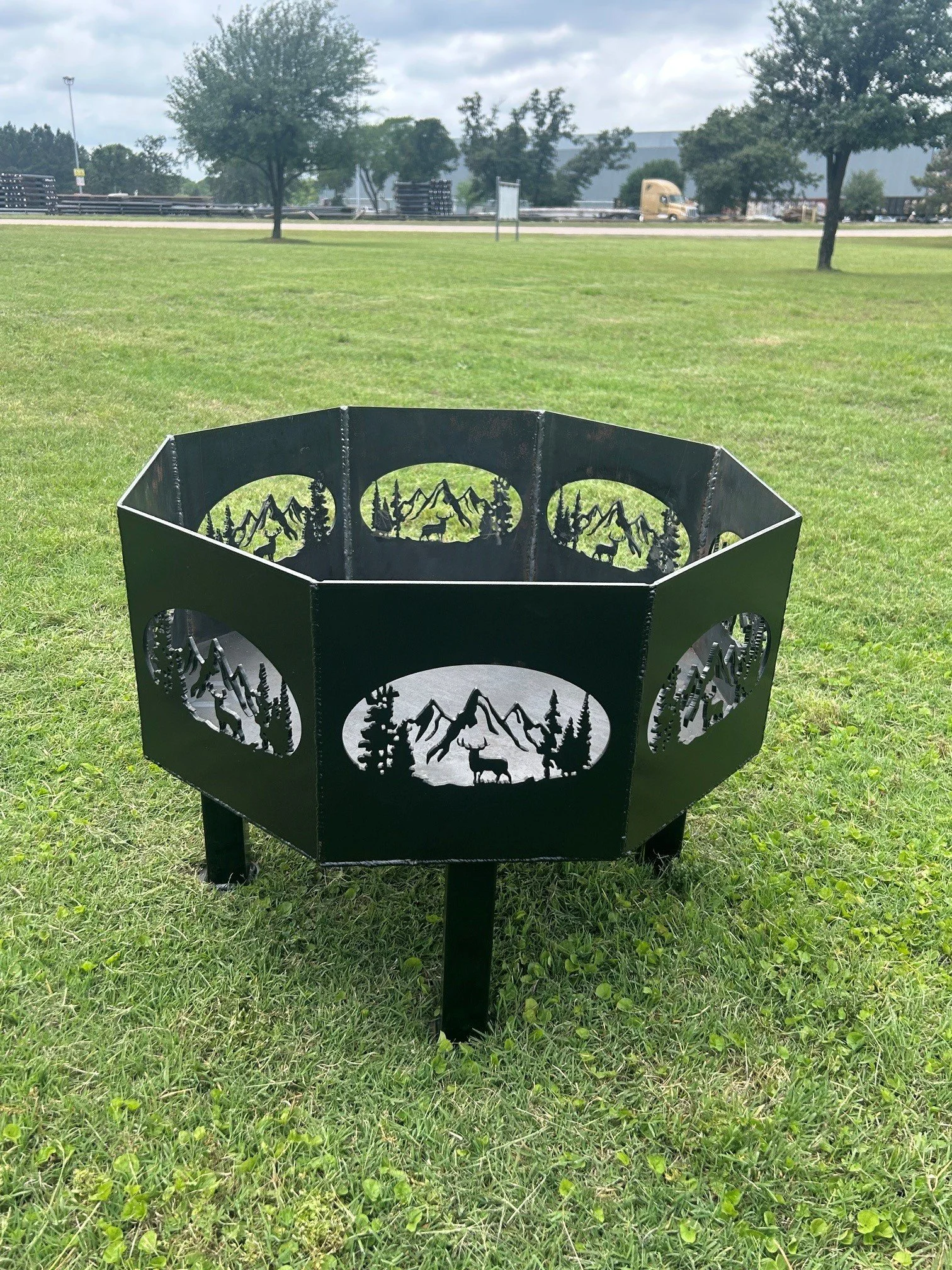 A decorative metal fire pit with mountain, forest, and elk scenic design, situated on a grassy area with trees, a road, and a truck in the background.