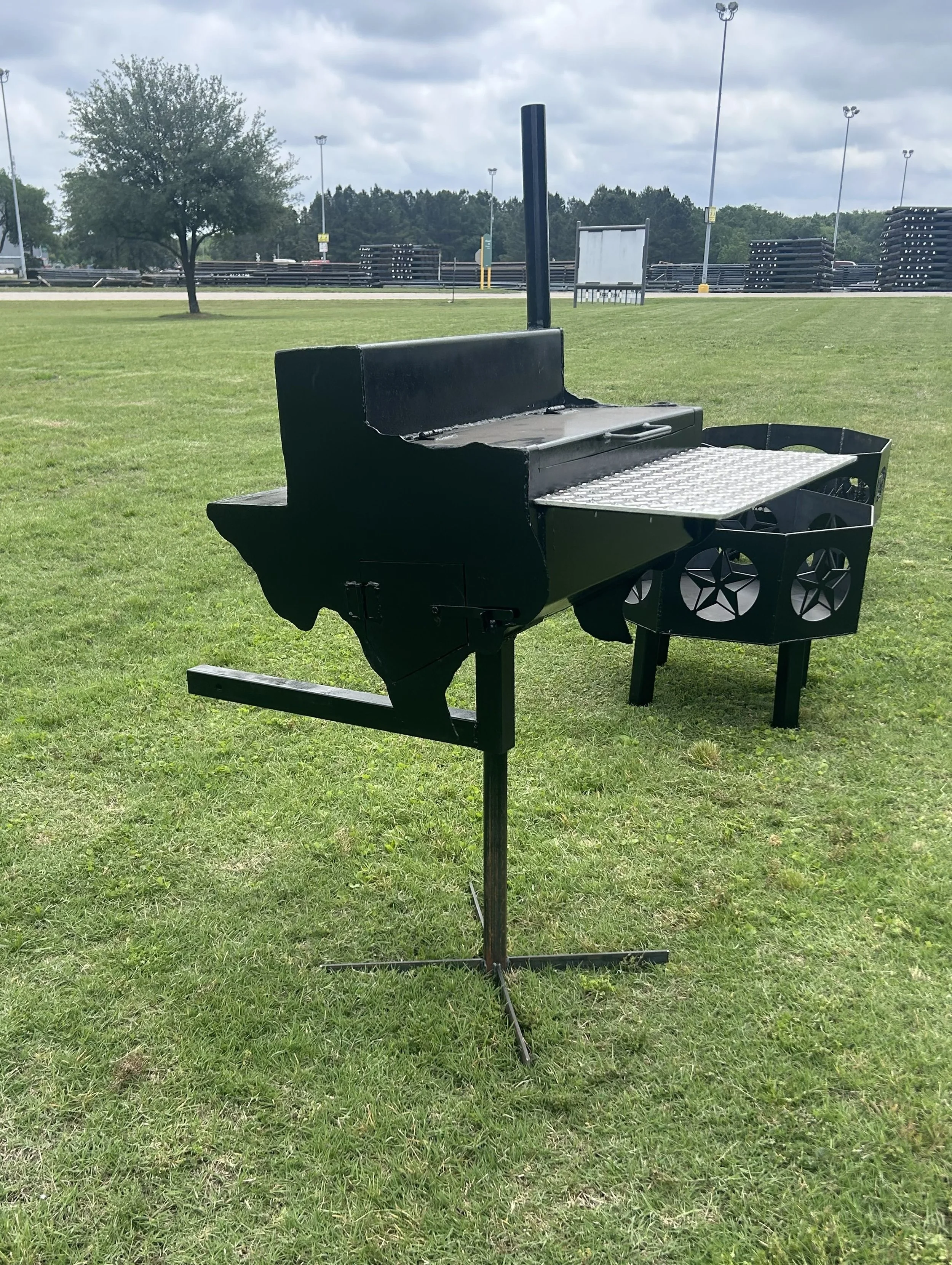A black barbecue grill with a metal cooking surface, situated on a grassy field with trees and a parking lot in the background, under a cloudy sky.