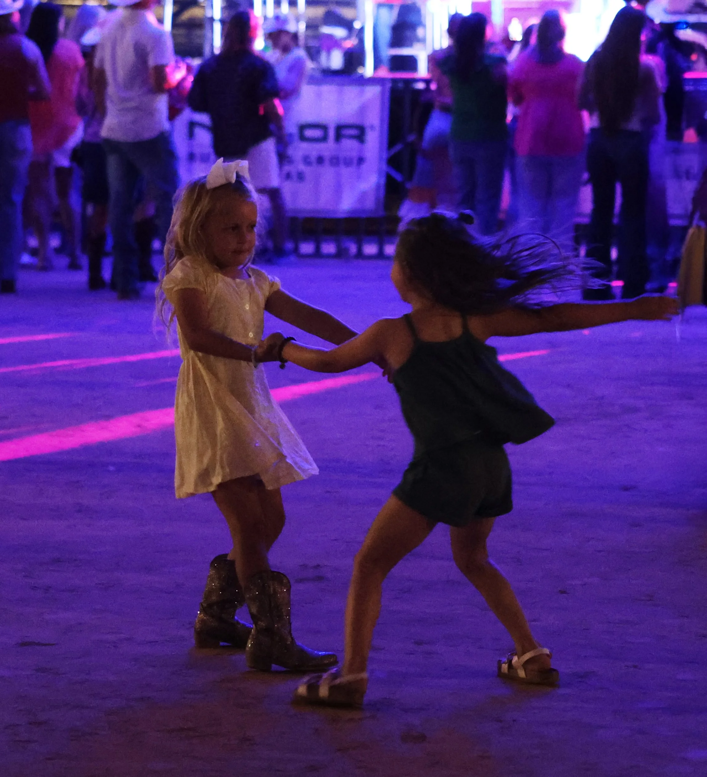 Two young girls dancing together in a dimly lit area with colorful neon lights, with other people standing and socializing in the background.