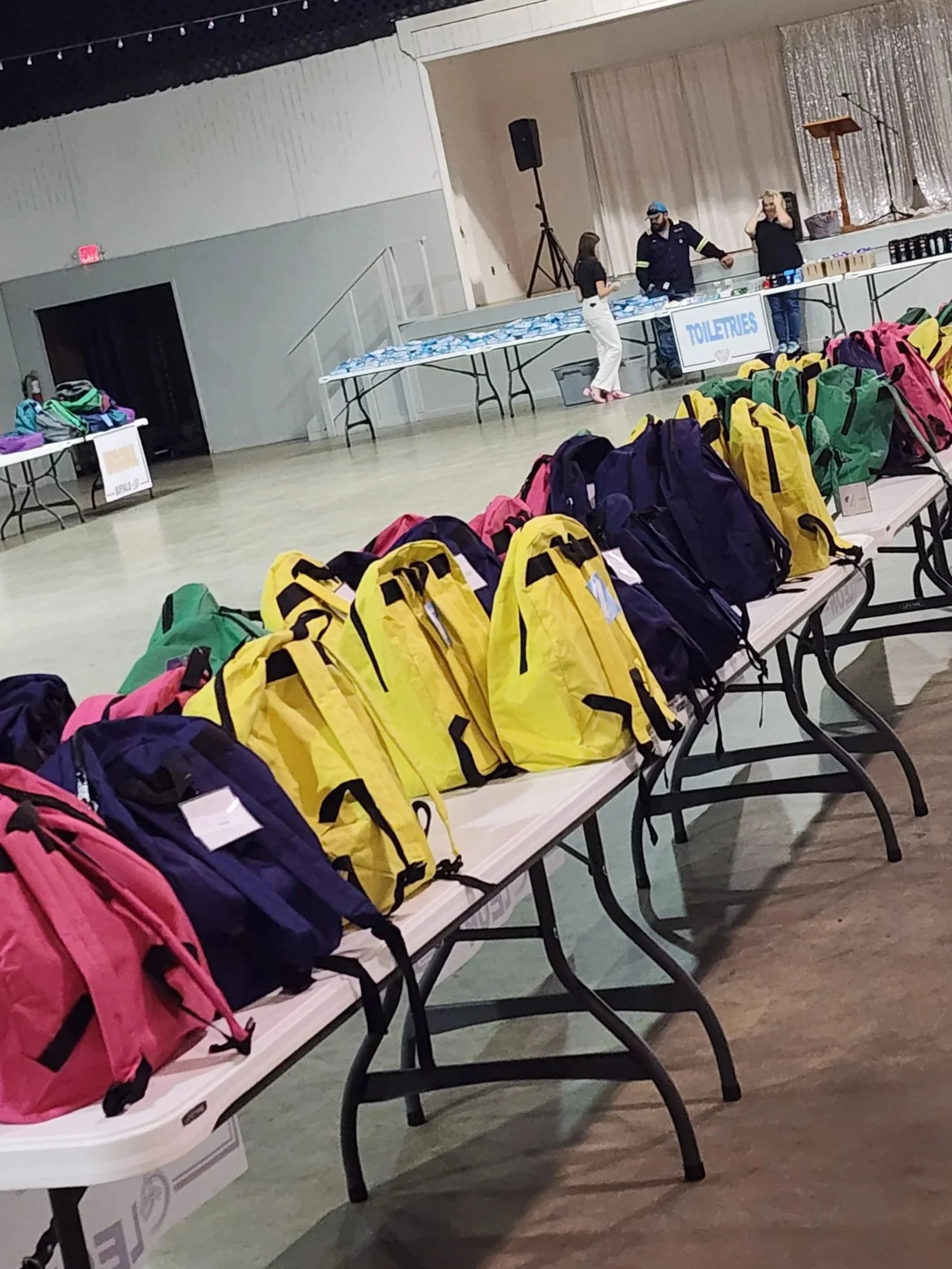 Indoor event with tables displaying backpacks of various colors, a sign that reads 'SENTELLOUS,' and a stage with people talking and standing.
