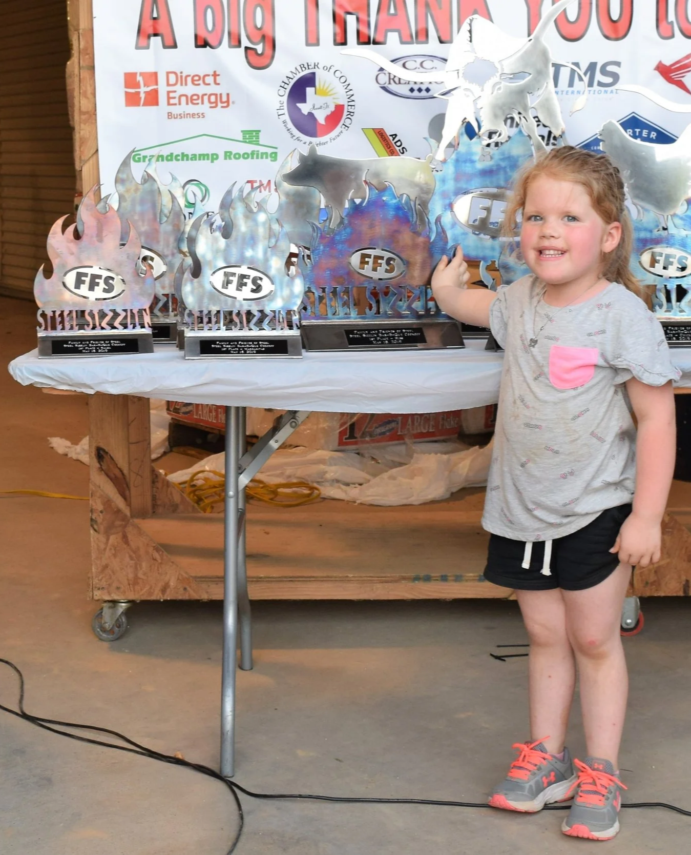 A young girl with curly red hair, grey shirt with pink pocket, and black shorts stands in front of a table with multiple awards. The awards are flame-shaped trophies with the FFS logo, displayed on a table covered with a white cloth, against a background with signs and banners.
