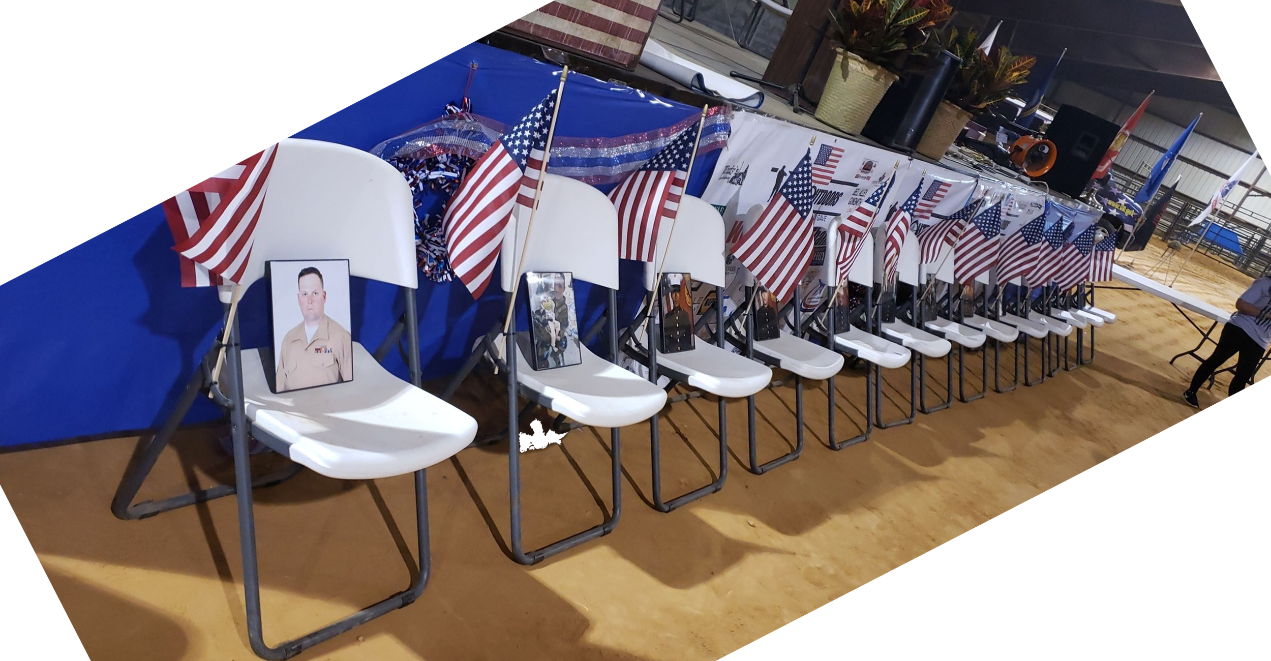Inside a venue decorated with American flags, white chairs are set up in a row with photographs of individuals in uniform placed on some of the seats, suggesting a patriotic or memorial event. A table with holiday decorations is in the background.