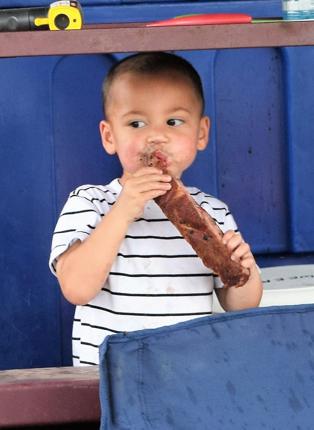 A young boy in a white and black striped shirt eating a large piece of cooked meat, with a blue background behind him.