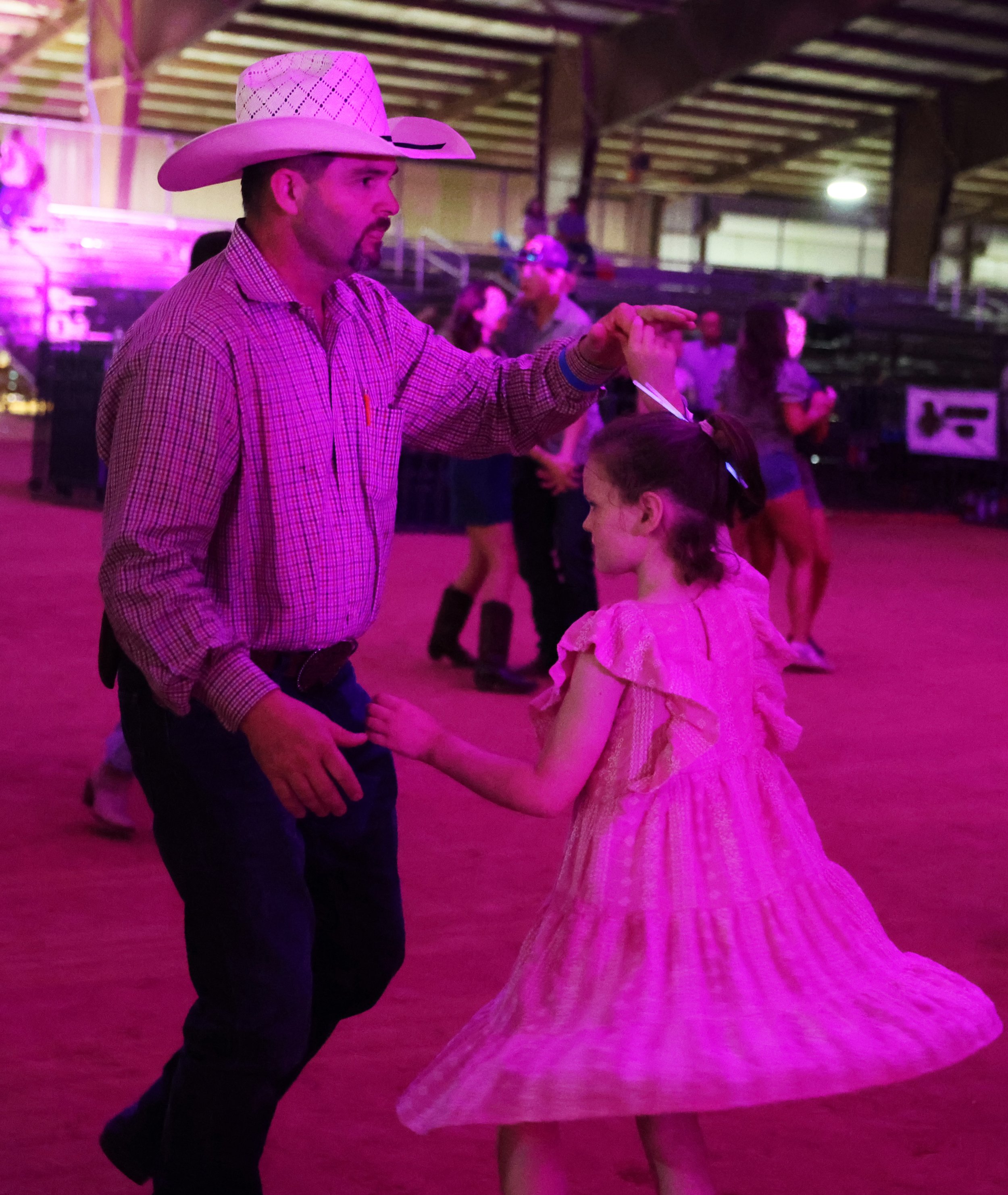 A man and a young girl dancing together in a dimly lit indoor space with pink and purple lighting. The man is wearing a cowboy hat and a checkered shirt, while the girl is in a light-colored dress.