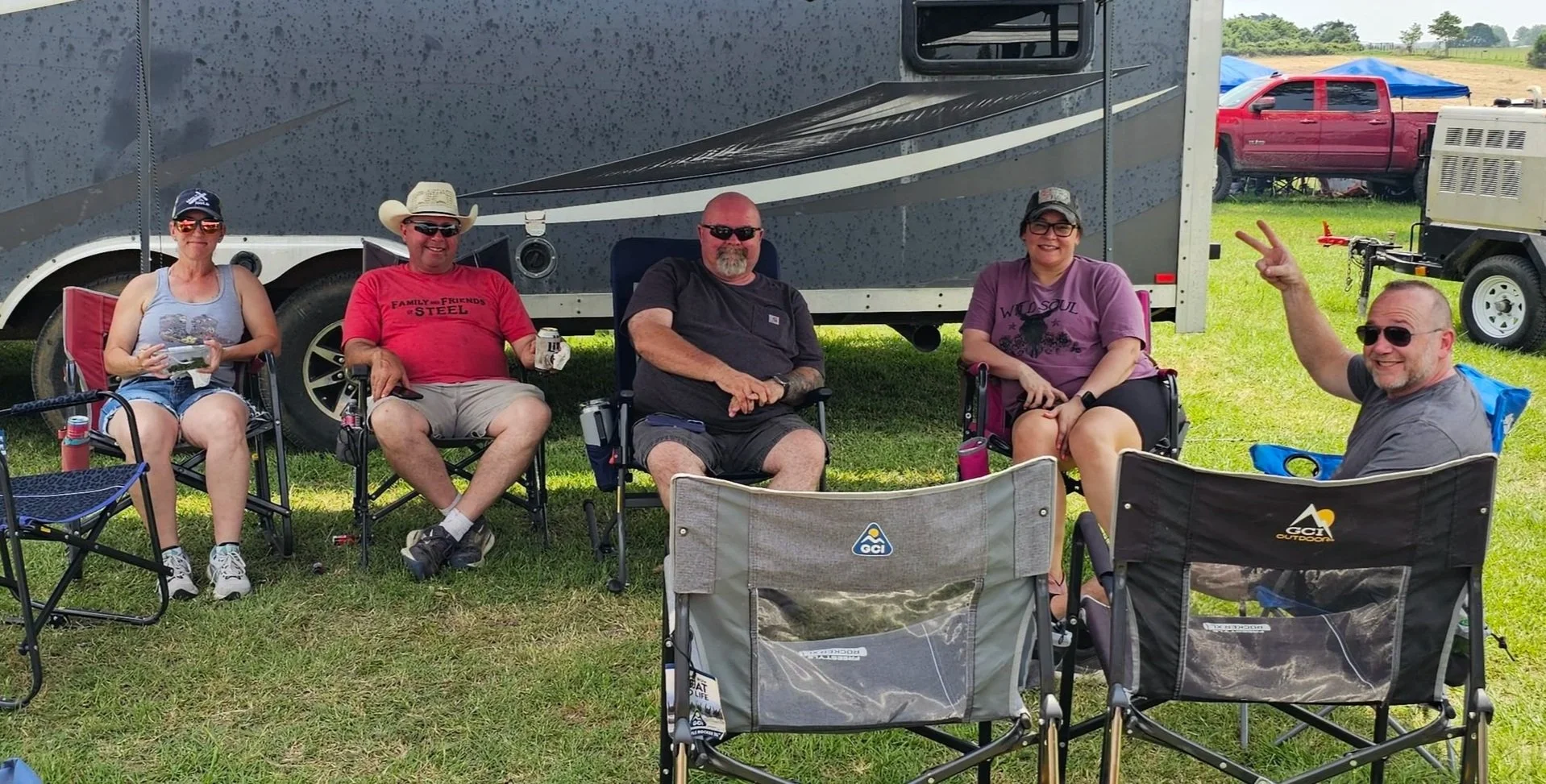 Group of five friends sitting outdoors in chairs near a camper trailer, with a red truck and generator in the background, during a sunny day.
