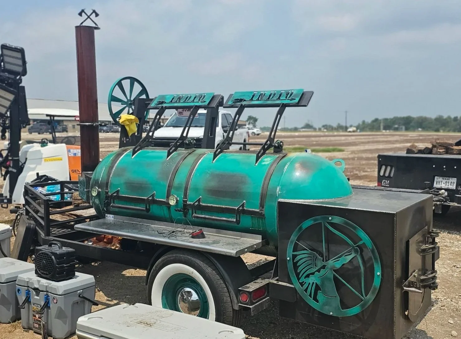 A green barbecue smoker on a trailer with a sunset or sunrise sky in the background, surrounded by equipment and vehicles in an open field.