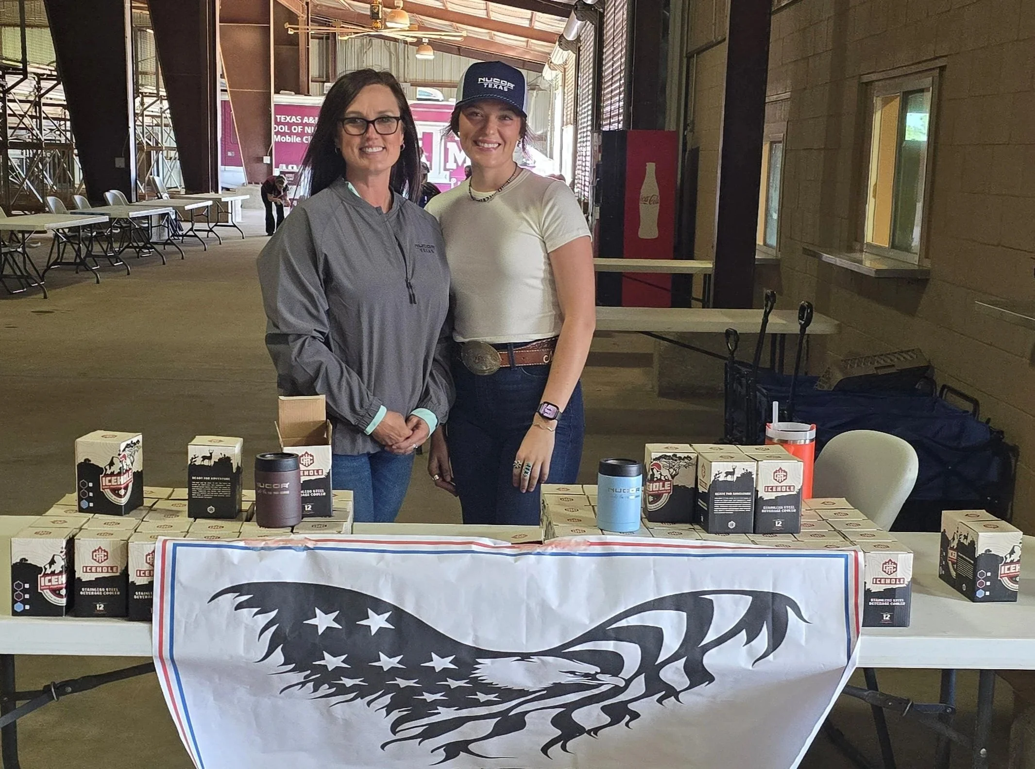 Two women standing behind a table with various products, including mugs and boxed items, with a large American flag-themed banner on the front of the table. They are inside a building with wooden walls and a high ceiling. One woman wears glasses and a gray jacket, while the other wears a cap, white t-shirt, and jewelry, both smiling at the camera.