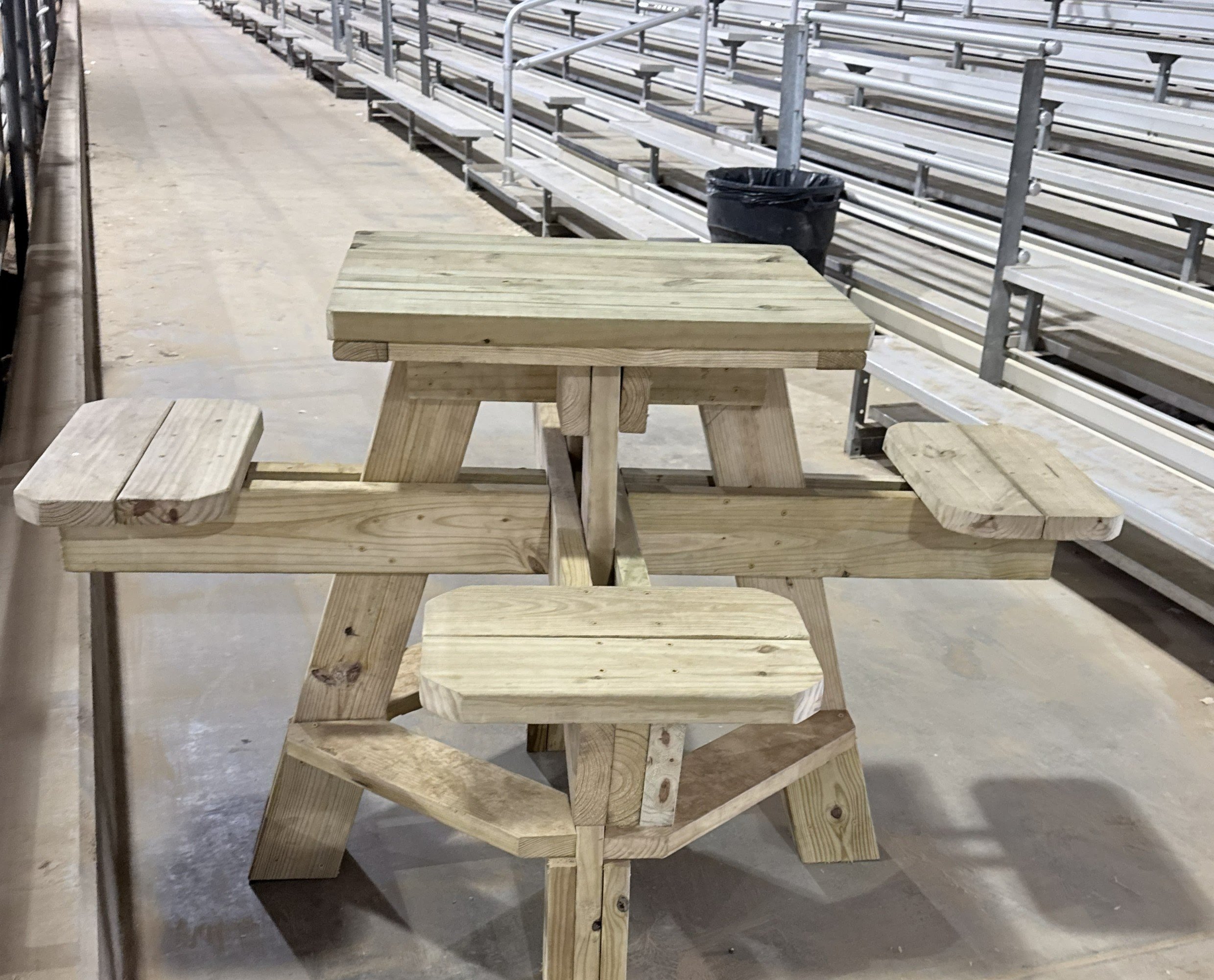 Wooden picnic table and benches next to aluminum bleachers and a trash can inside a stadium or sports field.