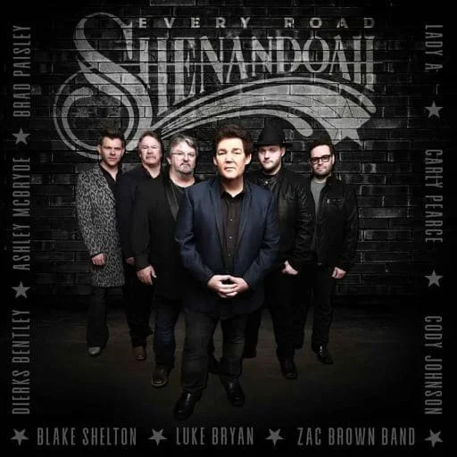 Five men standing in front of a black brick wall with "STAND AND DARE" written on it. The man in the center wears a dark blazer and looks directly at the camera, with the others standing behind him. Names of band members and song titles are written along the sides of the wall.