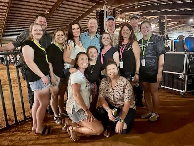 Group of 12 smiling people, mostly women, posing together indoors at an event with a wooden ceiling and metal fences, some wearing lanyards.