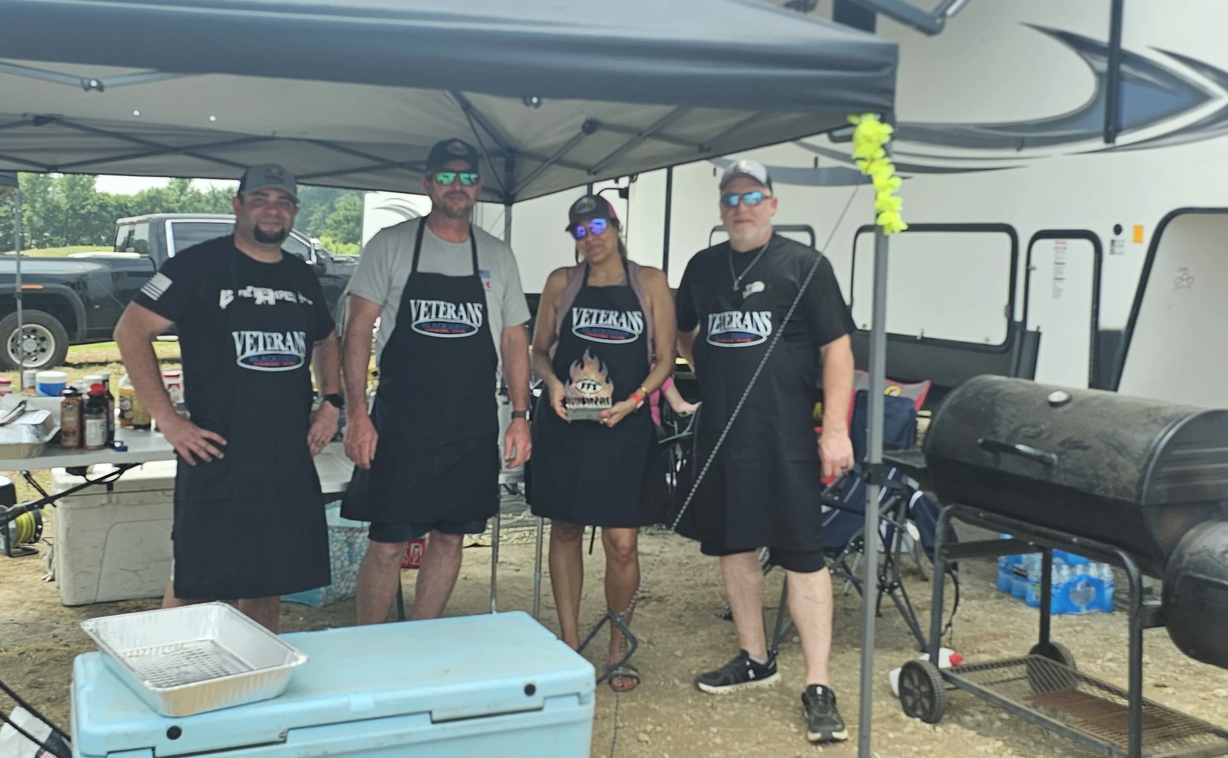 Four people standing under a canopy at an outdoor event, wearing shirts and aprons with 'VETERANS' printed on them. There are tables with food and drink, and a grill to the right. The background shows vehicles and trees.