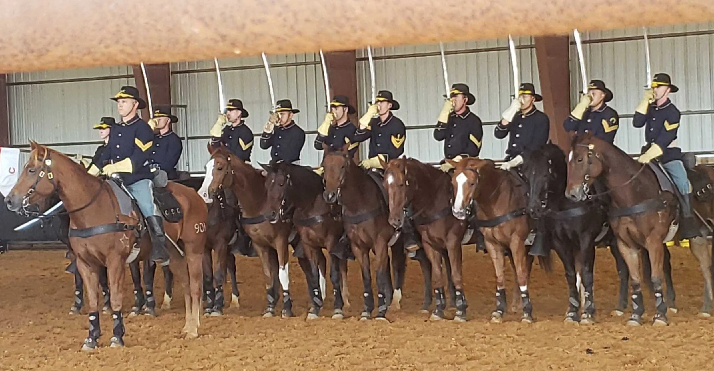 Group of riders in uniform on horseback inside an indoor arena, holding swords upright during a ceremony or parade.