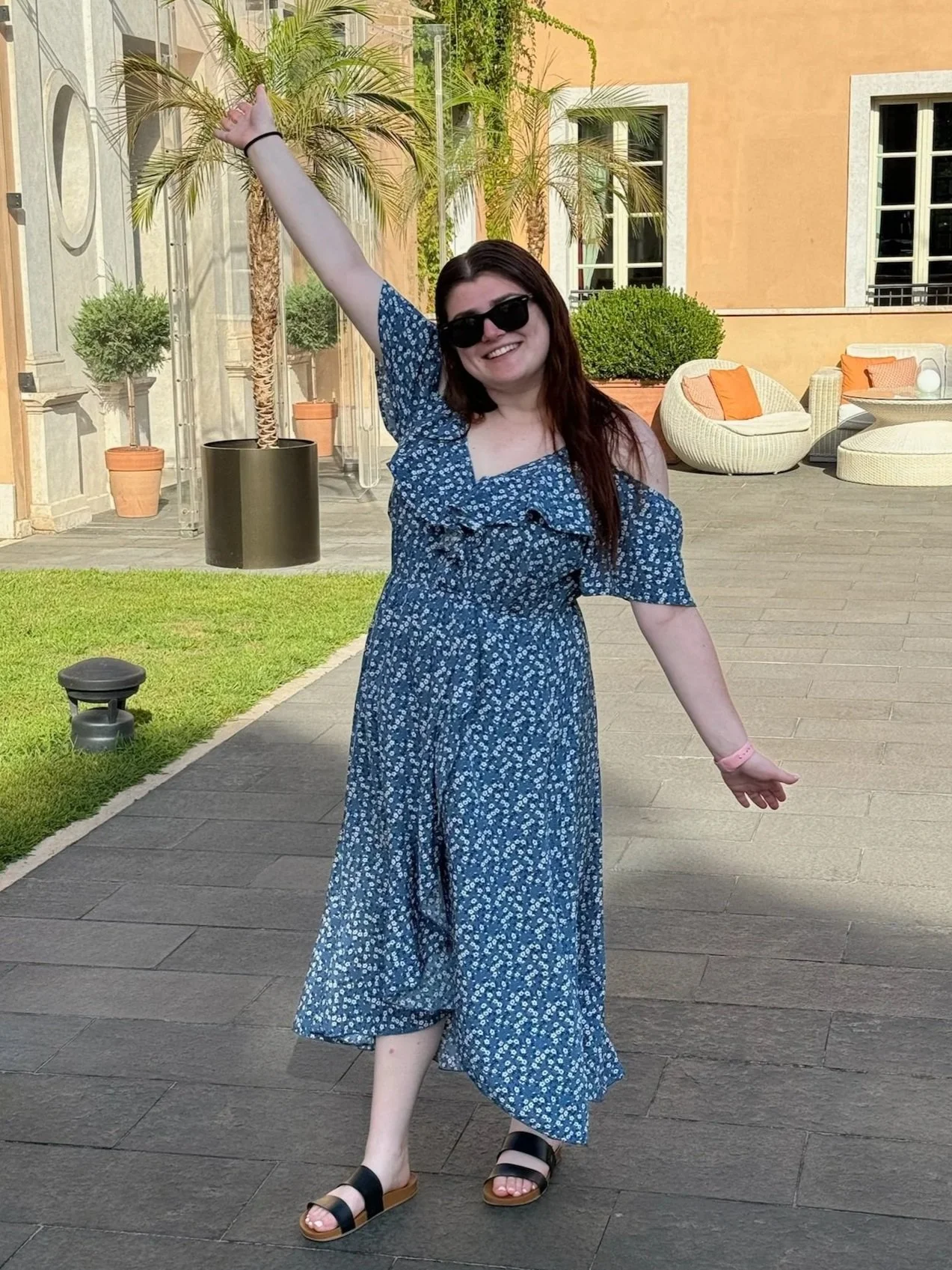 A young woman with long dark hair, wearing sunglasses and a blue floral dress, standing outdoors on a paved patio with her arms extended. Behind her are potted plants, including palm trees, and modern outdoor furniture in a sunny setting.