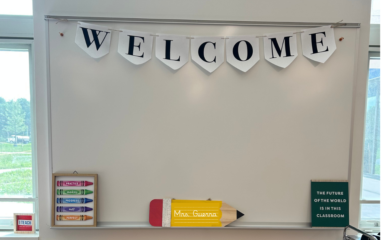 Classroom with a whiteboard decorated with a 'Welcome' banner, a small framed sign saying 'I Teach', a large pencil-shaped sign labeled 'Mrs. Guerra', and a green sign stating 'The future of the world is in this classroom'.