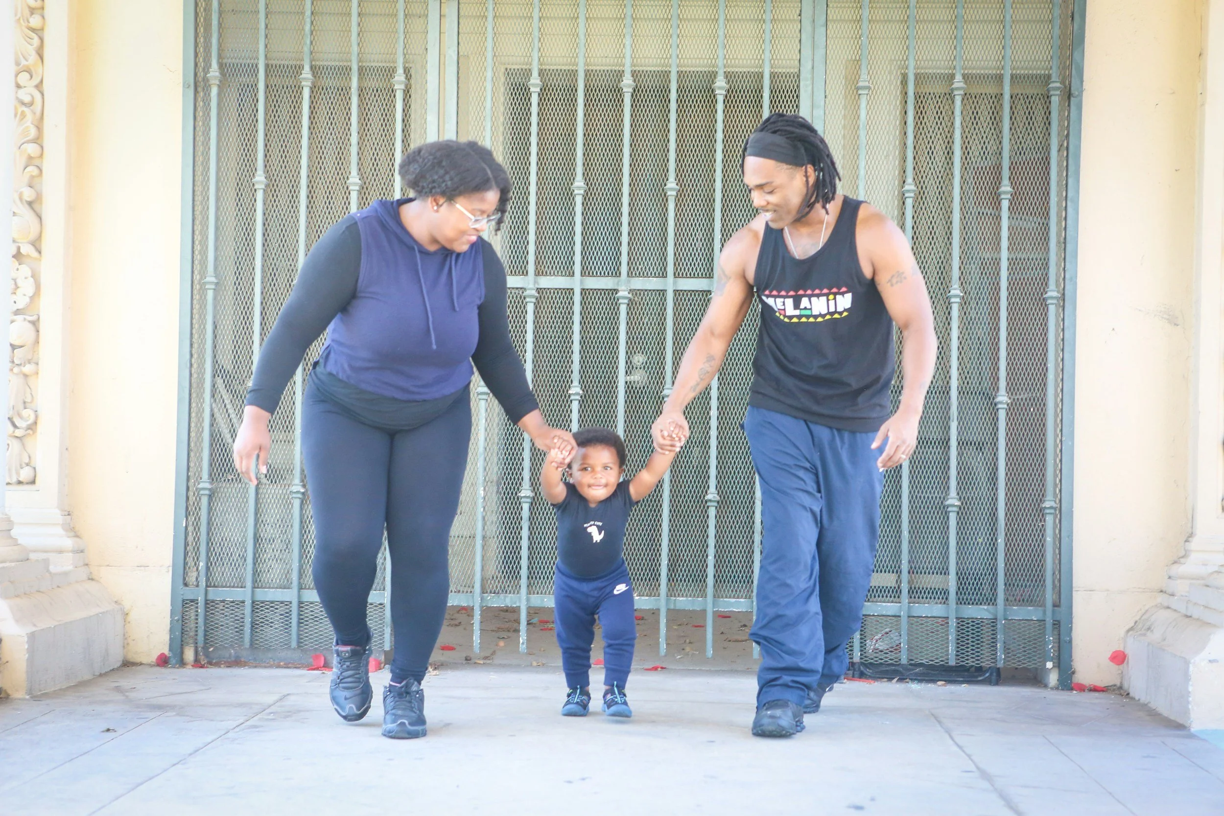 A family of three, a woman, a man, and a small child, walking together outside on a sidewalk in front of a gate. The woman is wearing glasses, a blue and black long-sleeve shirt, and black pants. The man is dressed in a black tank top with colorful text and dark blue pants. The child is holding both of their hands and is wearing a dark shirt with a white elephant graphic, dark pants, and sneakers. They all appear to be smiling and enjoying each other's company.