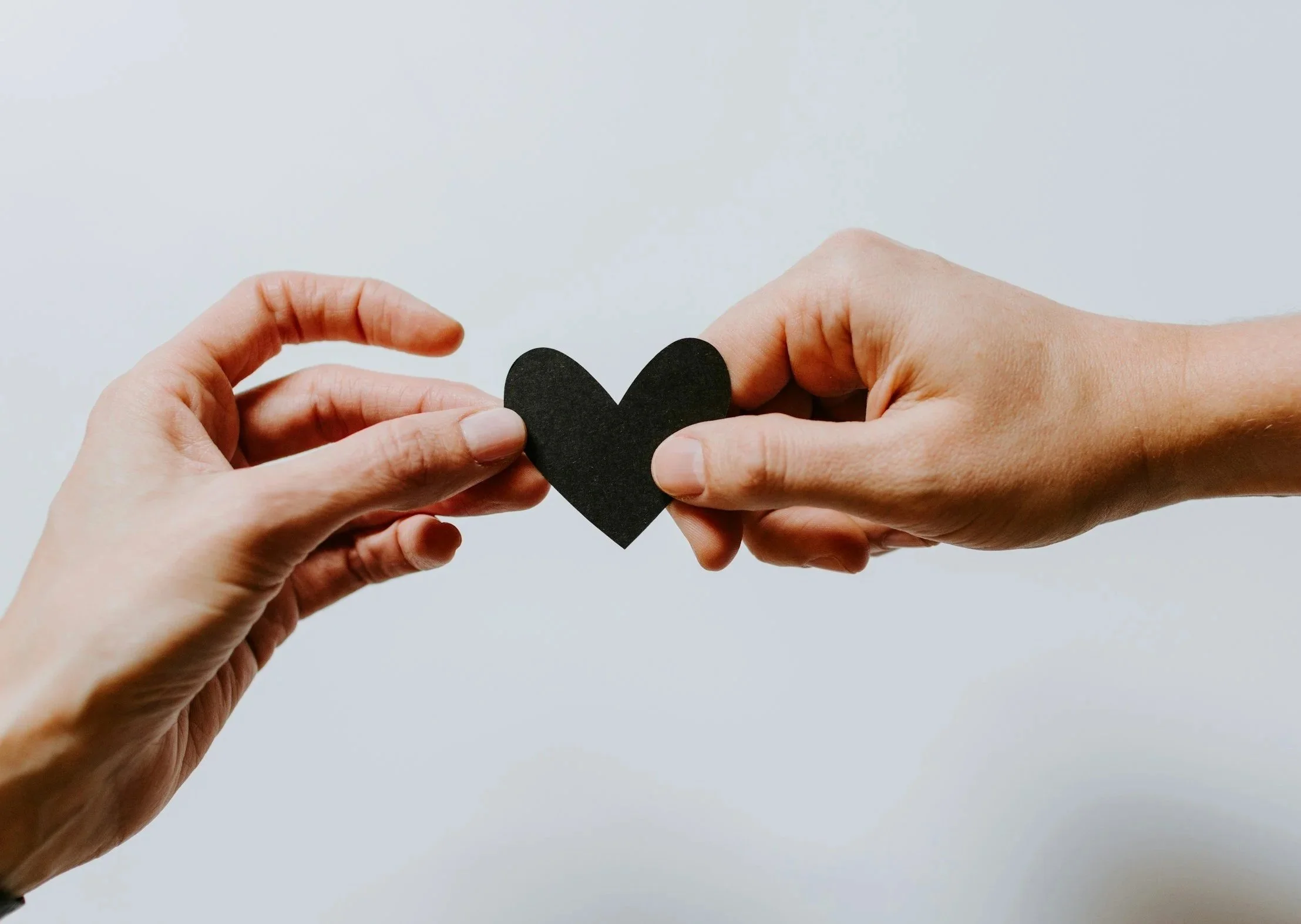 Two hands holding a small black heart cutout against a plain light grey background.