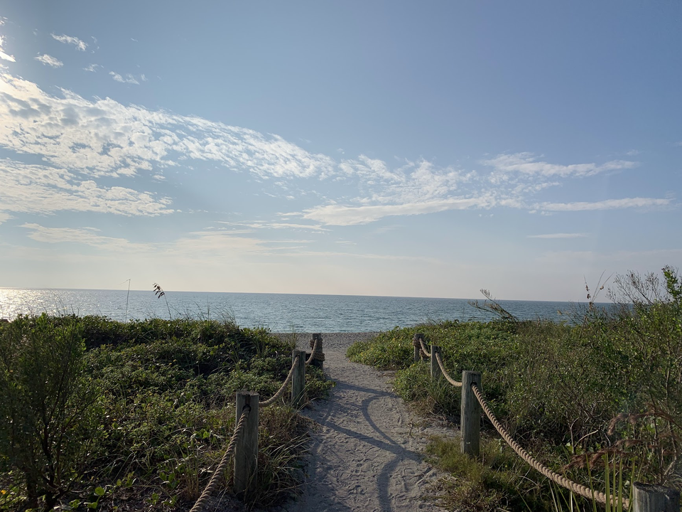 A sandy pathway leading to a beach, with green bushes on each side, and a calm ocean under a partly cloudy sky.