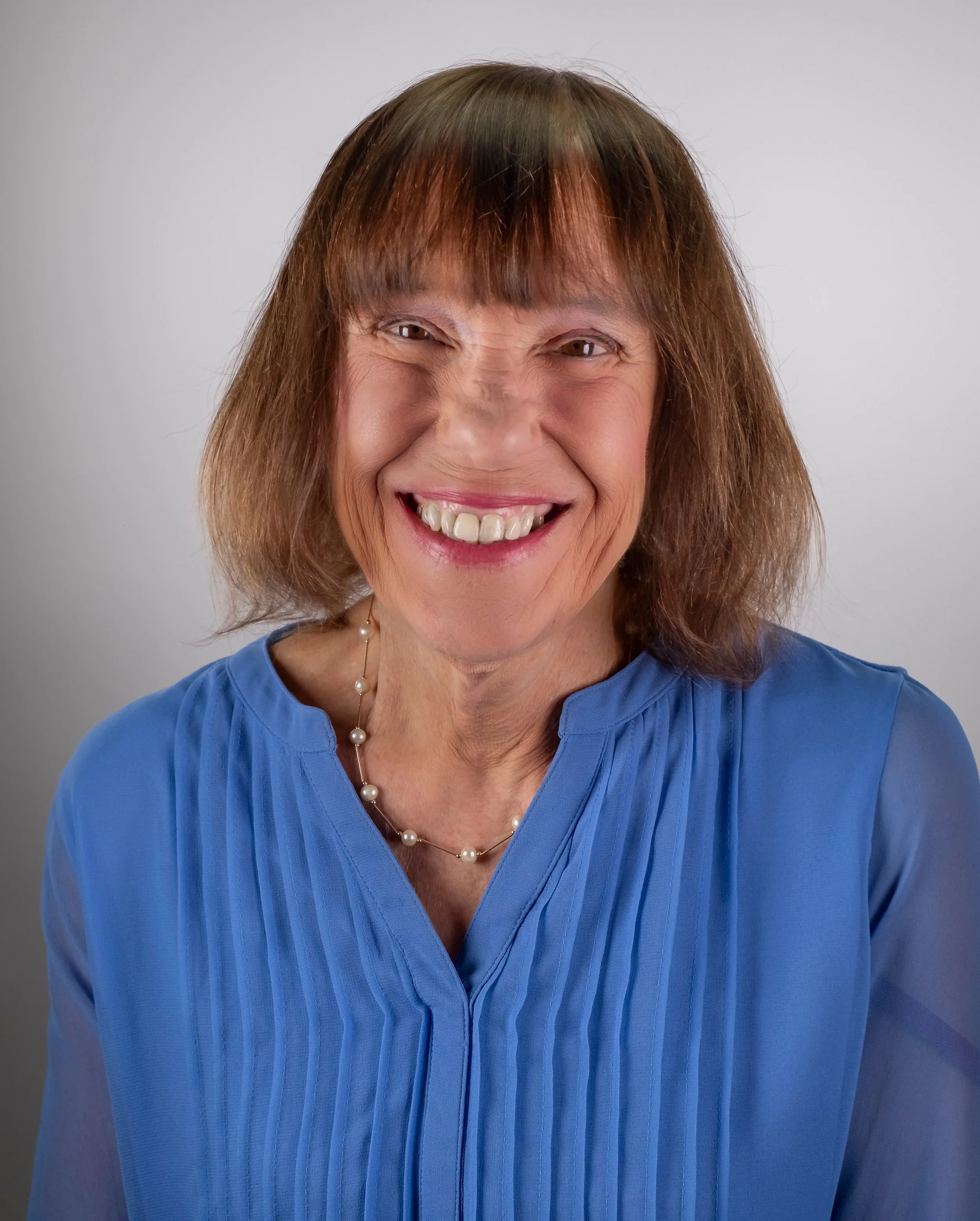 Close-up of an older woman with brown hair and bangs, smiling broadly, dressed in a blue top, wearing a pearl necklace against a light gray background.