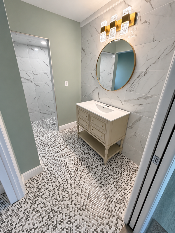 Completed bathroom renovation with sage green walls, marble-look tile, custom vanity and lighting, round mirror, and penny tile flooring