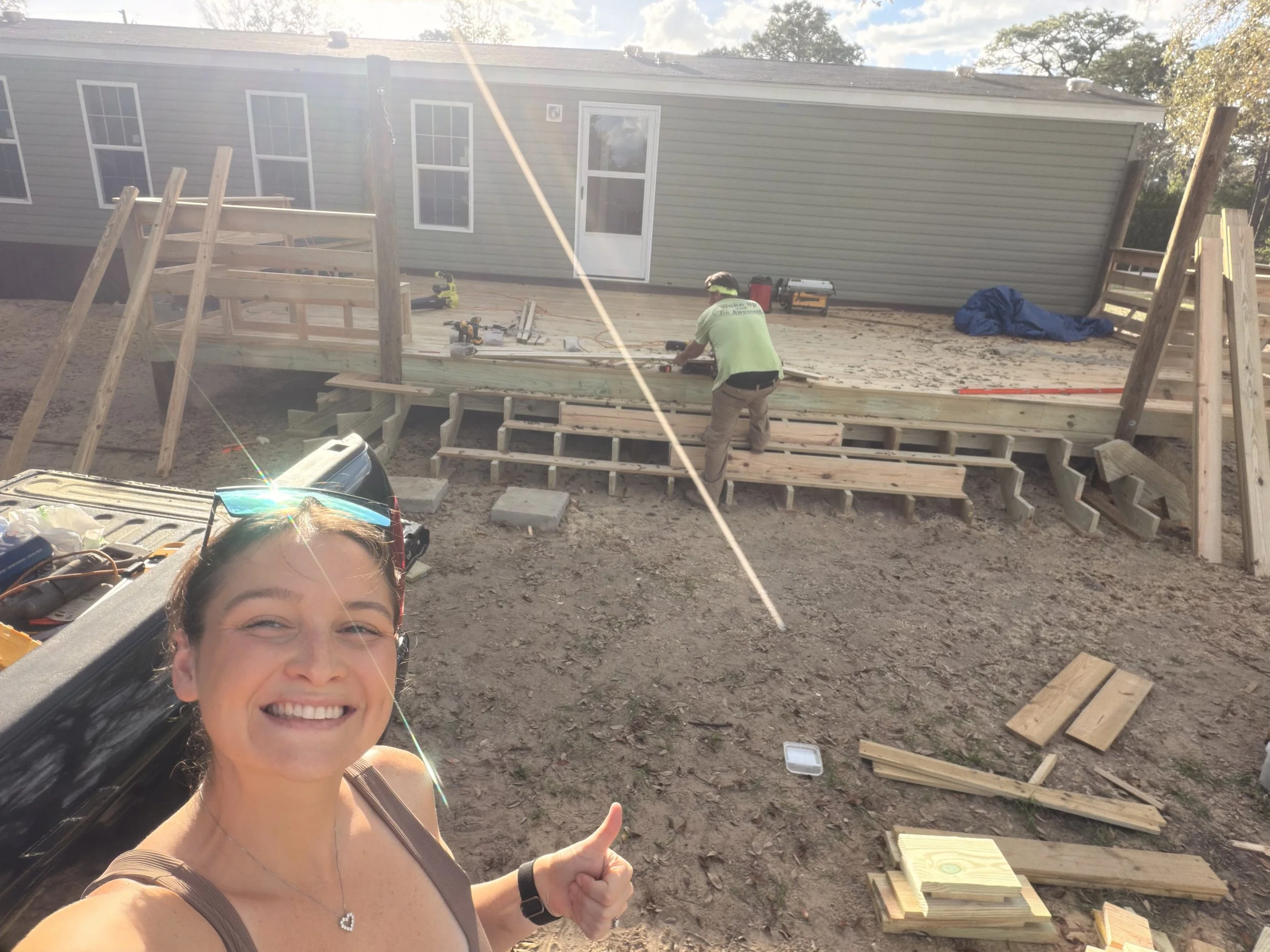 A woman giving a thumbs-up in front of a mobile home while a large raised wooden deck is being built, with deck framing, stairs, railings, and another worker visible