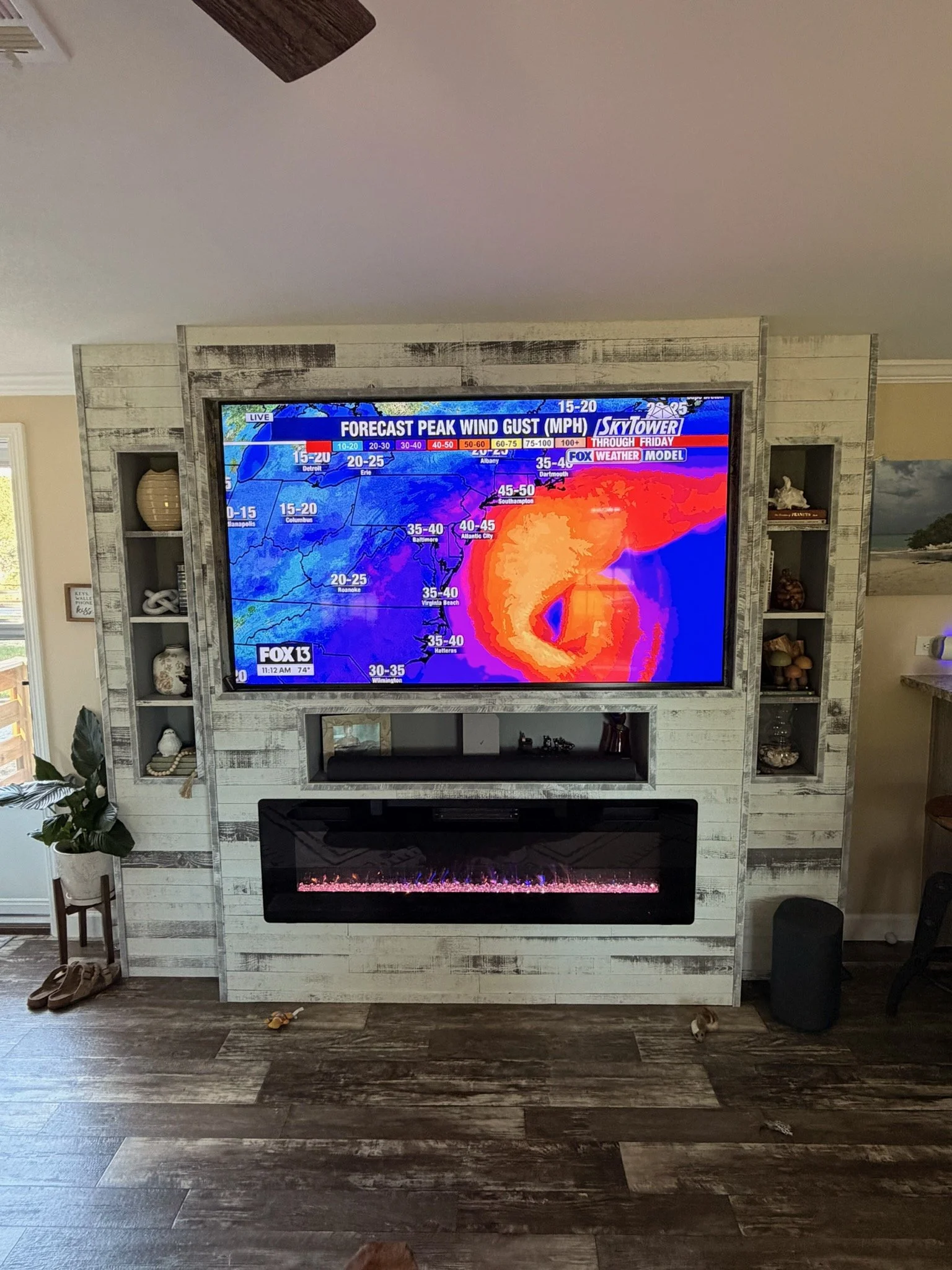 Completed custom living room entertainment center with a large TV, built-in electric fireplace, distressed wood finish, and built-in shelving on both sides.