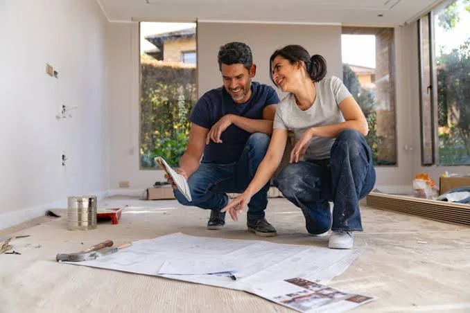A man and woman smiling and looking at a blueprint or large plan in an empty room with large windows and construction materials around.
