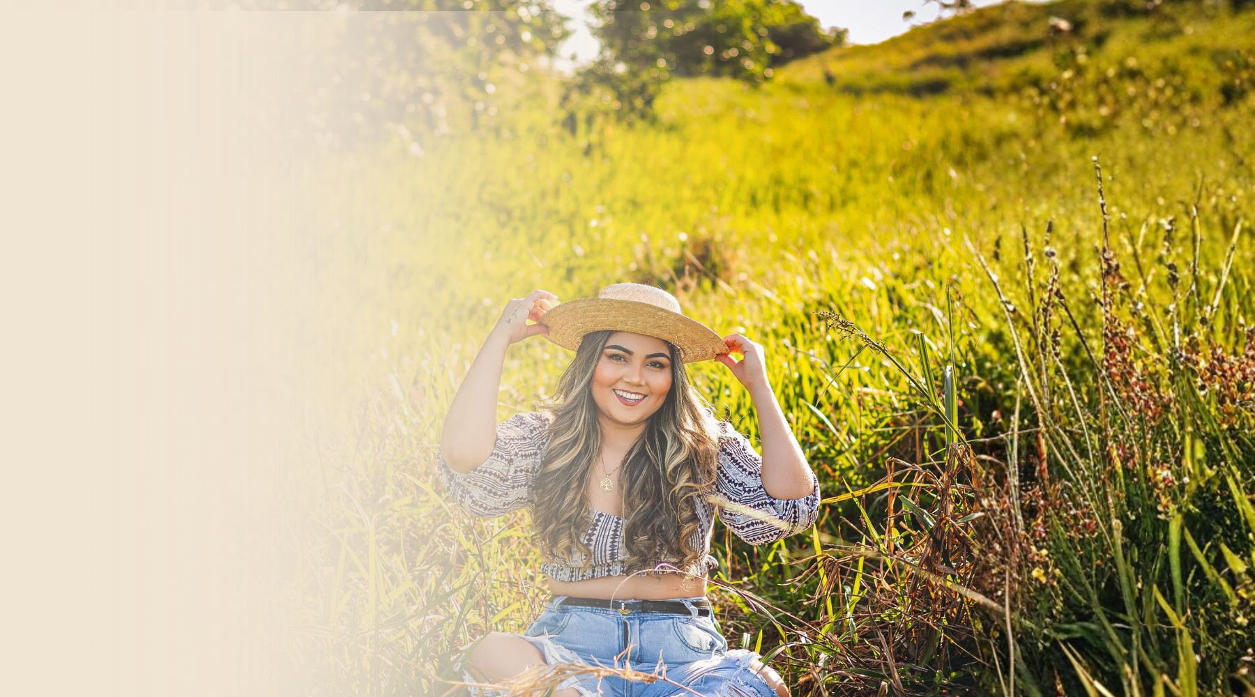 A young woman sitting in a field of tall grass, smiling and holding a wide-brimmed straw hat on her head, with sunlight and green foliage in the background.