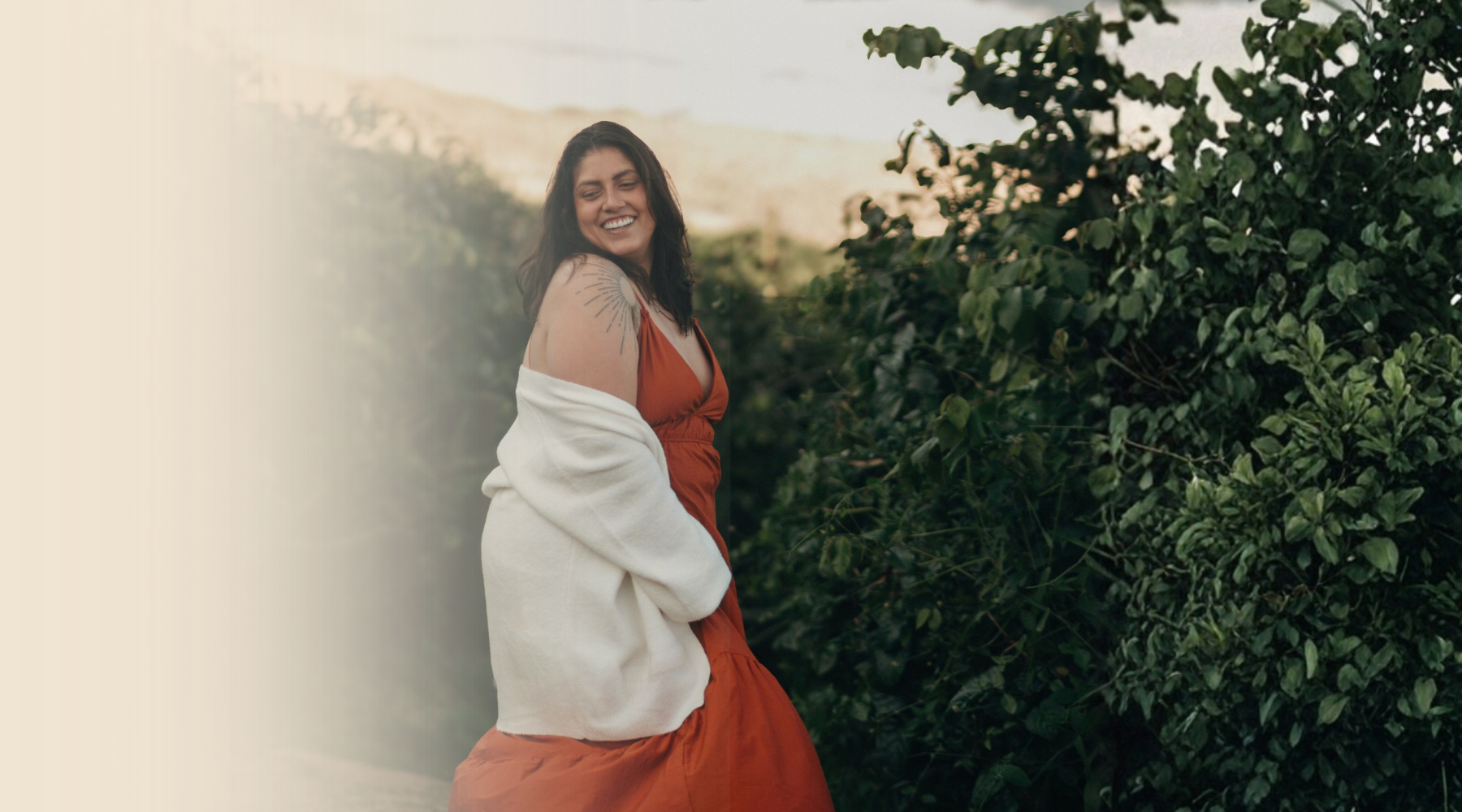 A smiling woman with dark hair, wearing a red dress and a white jacket, standing outdoors next to green bushes.