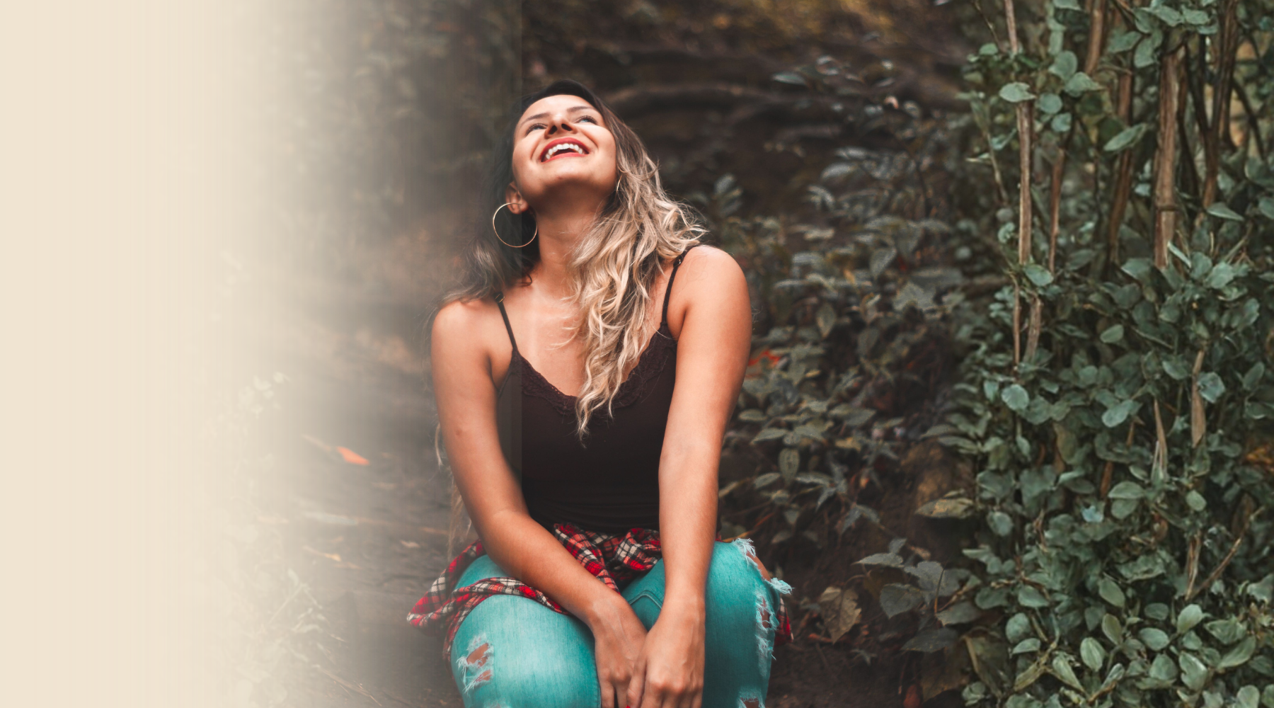 A woman with long wavy hair wearing a black tank top and ripped jeans, sitting outdoors near greenery, smiling and looking up.