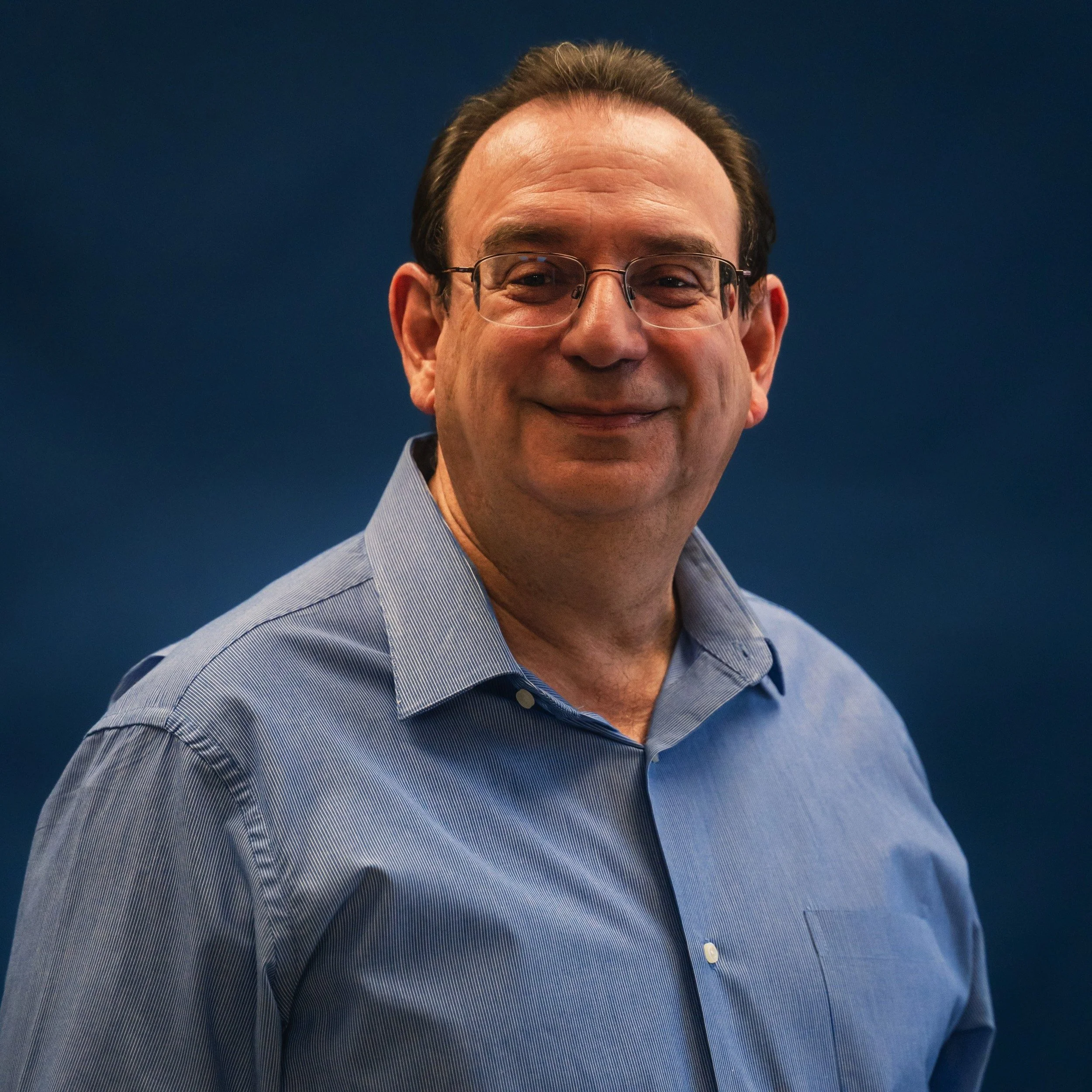 Close-up of a man wearing glasses and a blue striped shirt, smiling with a dark background.