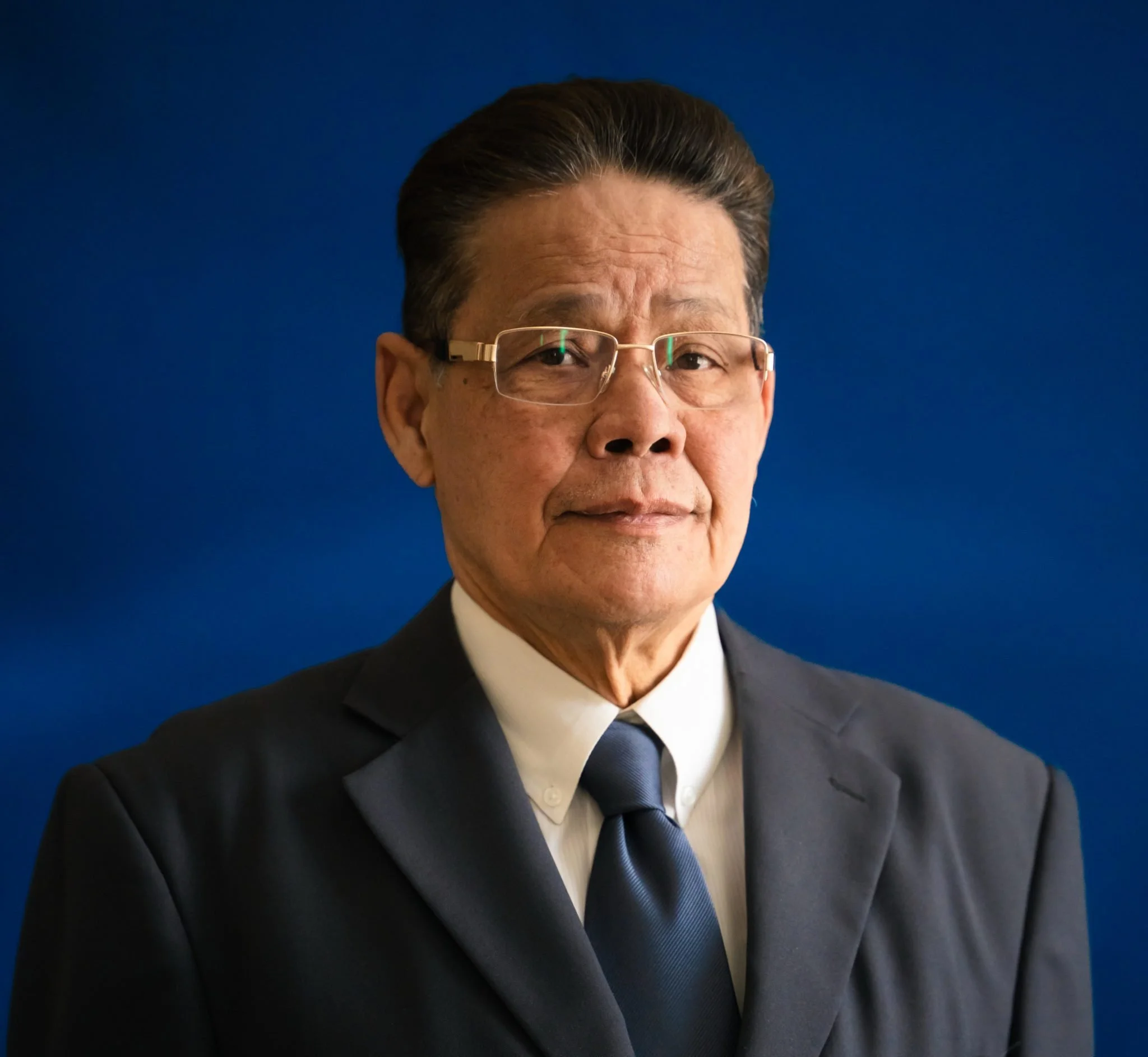 Close-up of a man wearing glasses, a tuxedo, a white shirt, and a black tie, with a dark blue background.