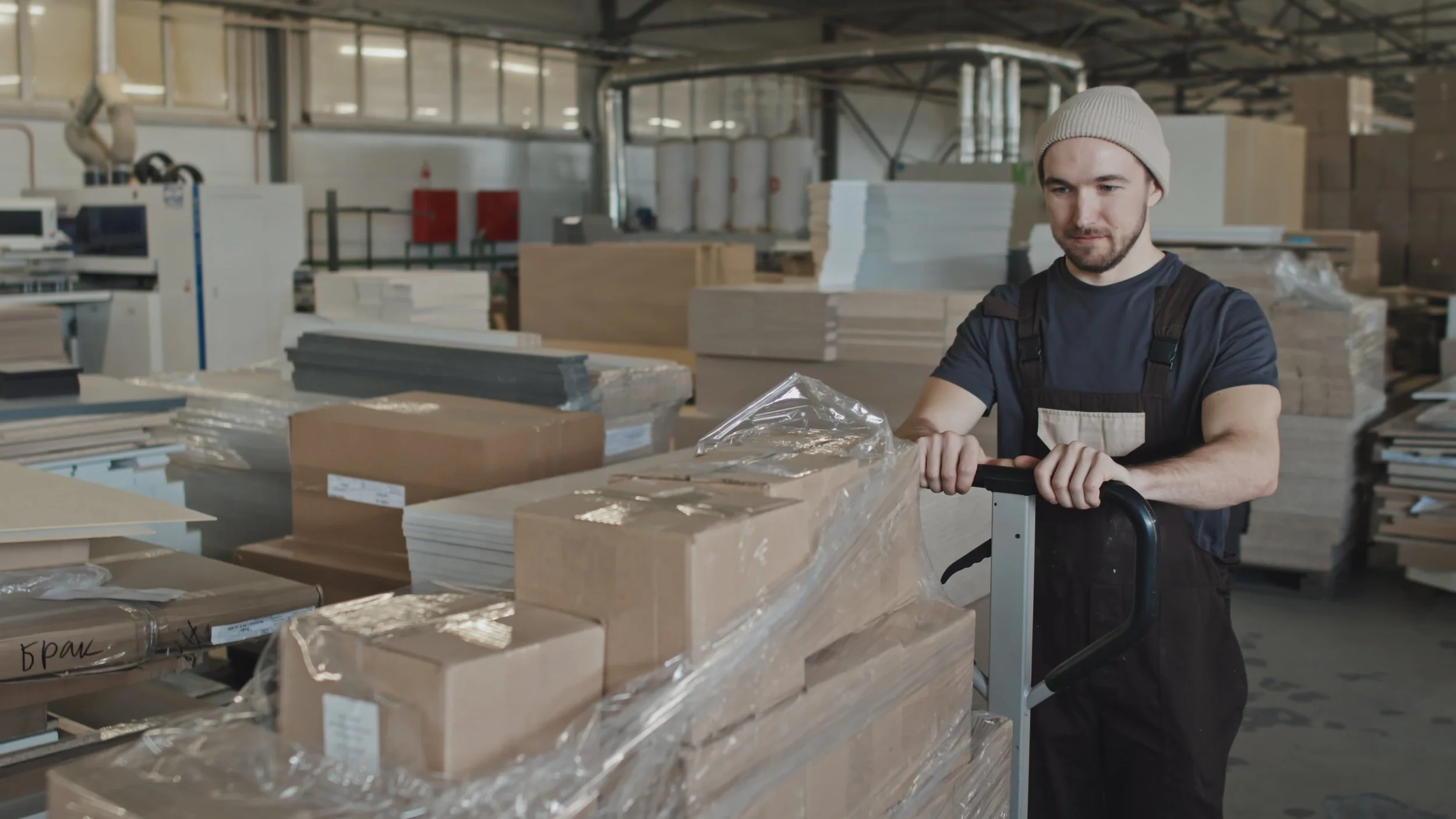 A man in a beanie and black work apron pushing a cart with stacked packages in a warehouse.
