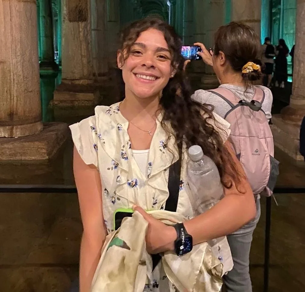 A young woman with curly brown hair and a bright smile poses for a photo inside a historic building, holding a water bottle and a camera bag, with other visitors in the background.