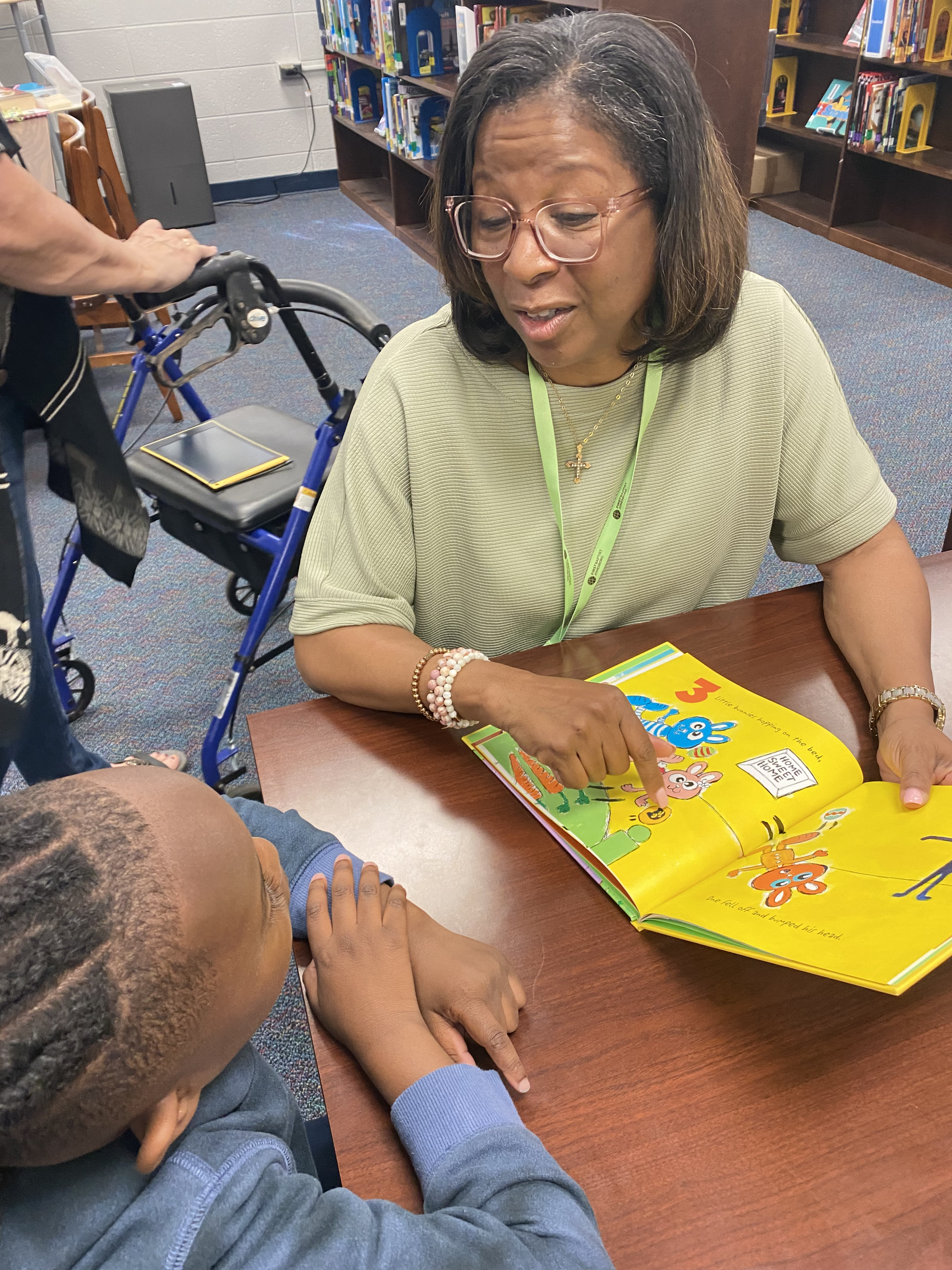 A woman with glasses reading a colorful children's book to a young boy with braids in a library. The woman is smiling and pointing at the book, which features cartoon characters and large text. The boy is resting his head on the table, listening attentively.