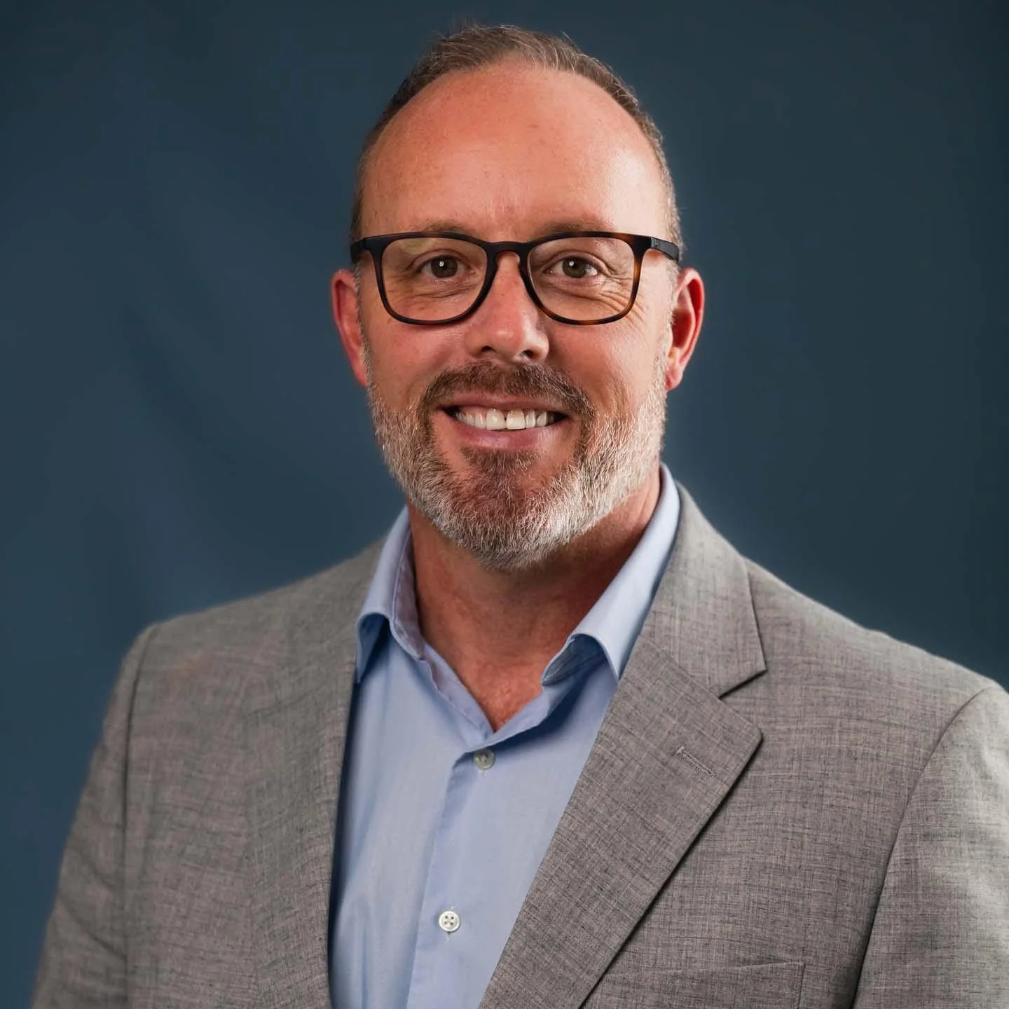 Headshot of a smiling man with glasses, a beard, wearing a gray blazer and light blue shirt, against a dark blue background.