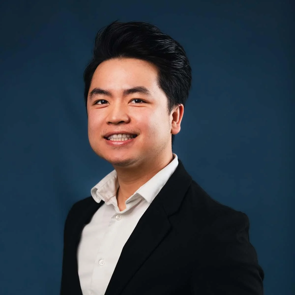 A young man with black hair, wearing a white shirt and black blazer, smiling against a dark blue background.