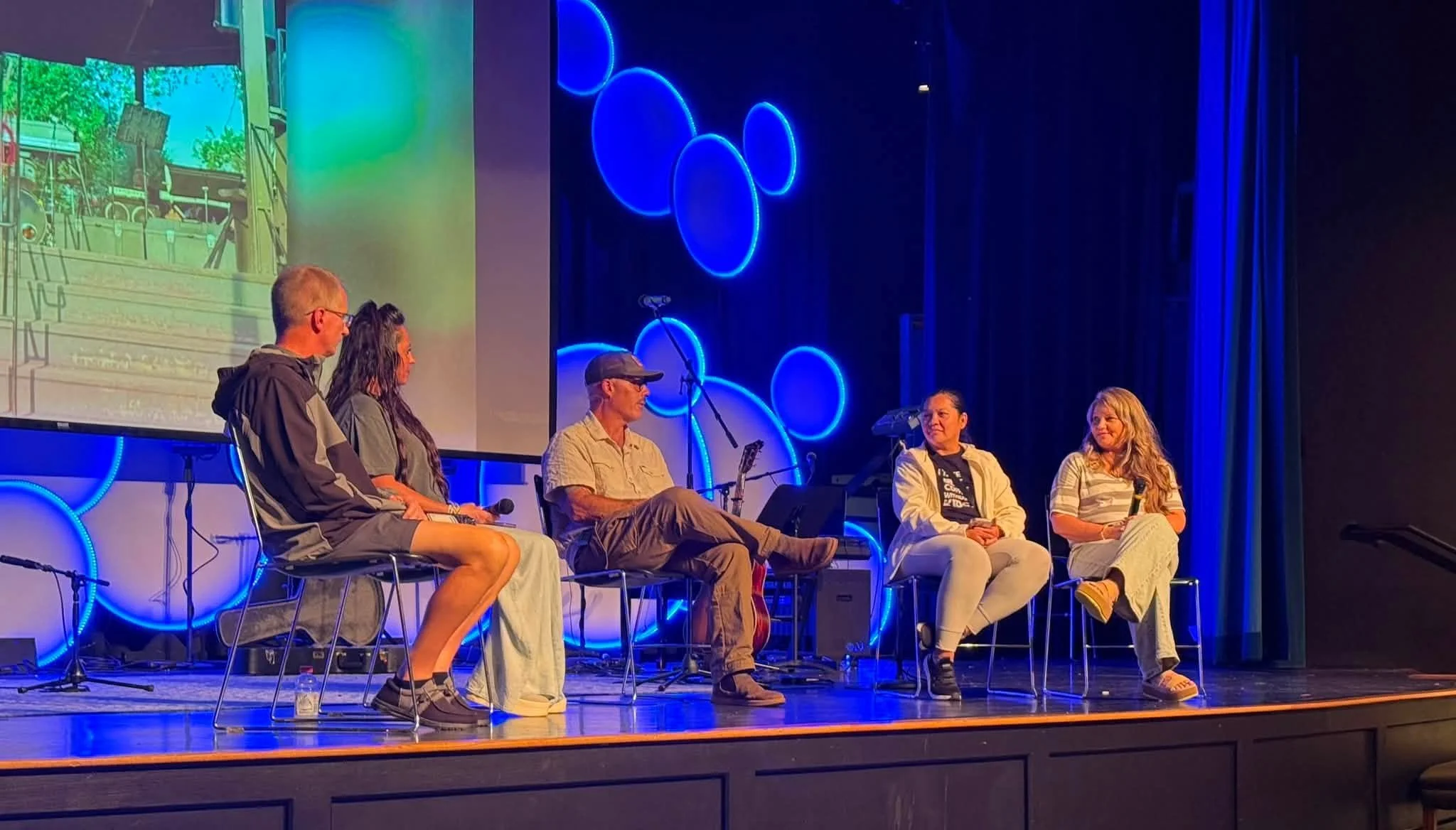 Six people sitting on a stage in front of a large screen, engaged in a panel discussion or presentation. The stage is decorated with blue neon circular lights.