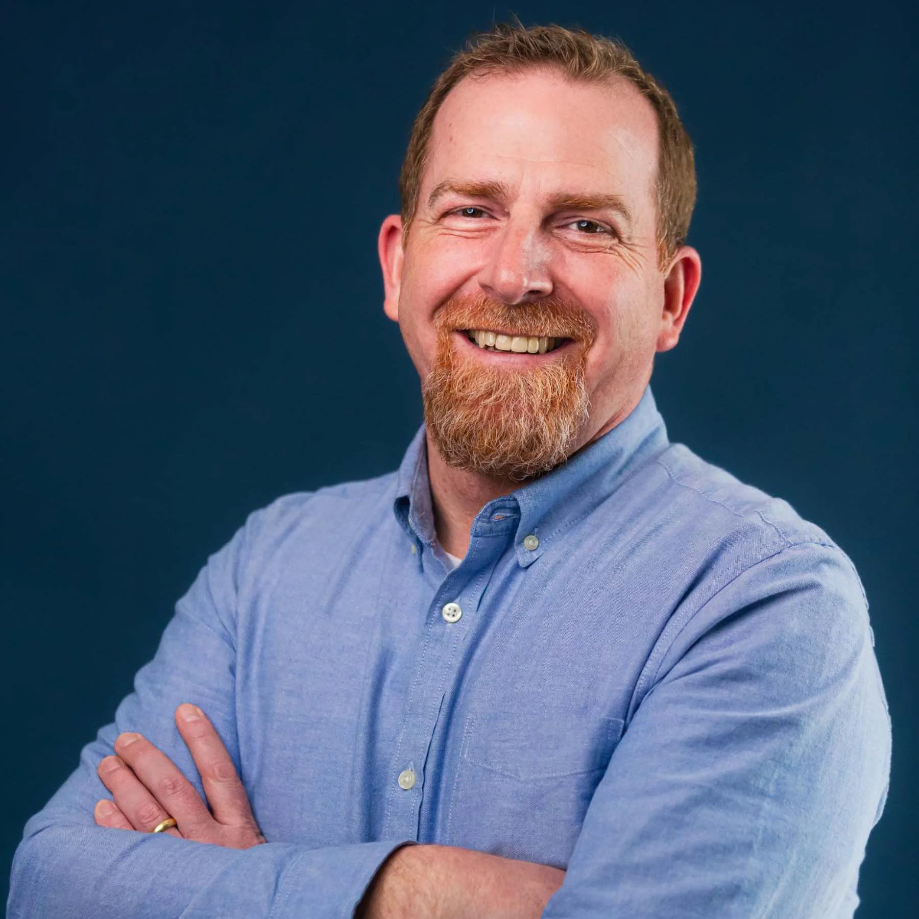 A smiling man with a red beard and short ginger hair, wearing a light blue button-up shirt, crossing his arms against a dark blue background.