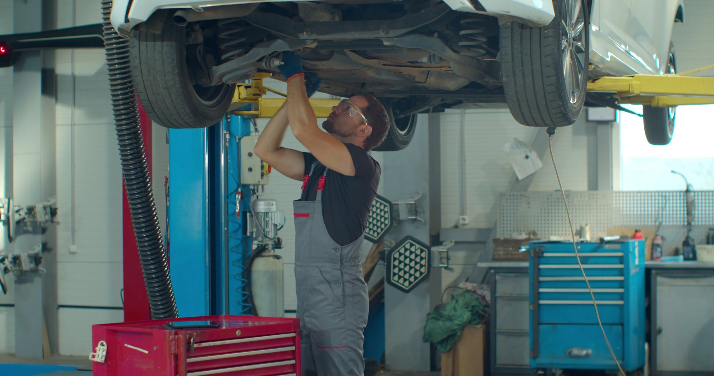 A mechanic working underneath an elevated white car in an auto repair shop.