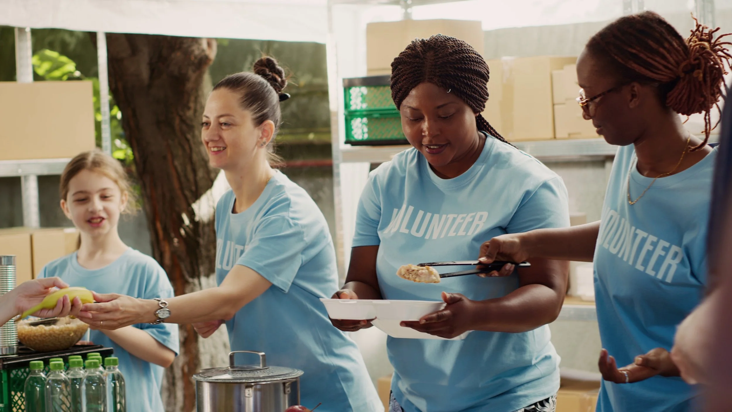 Group of diverse volunteers serving food at an outdoor event, wearing light blue shirts that say 'Volunteer'.