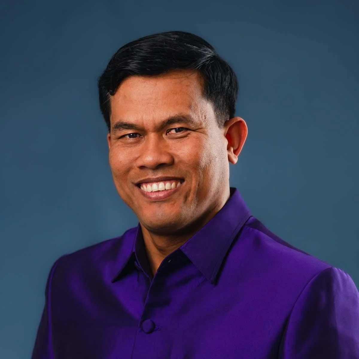 Portrait of a man in a purple shirt smiling against a blue background.