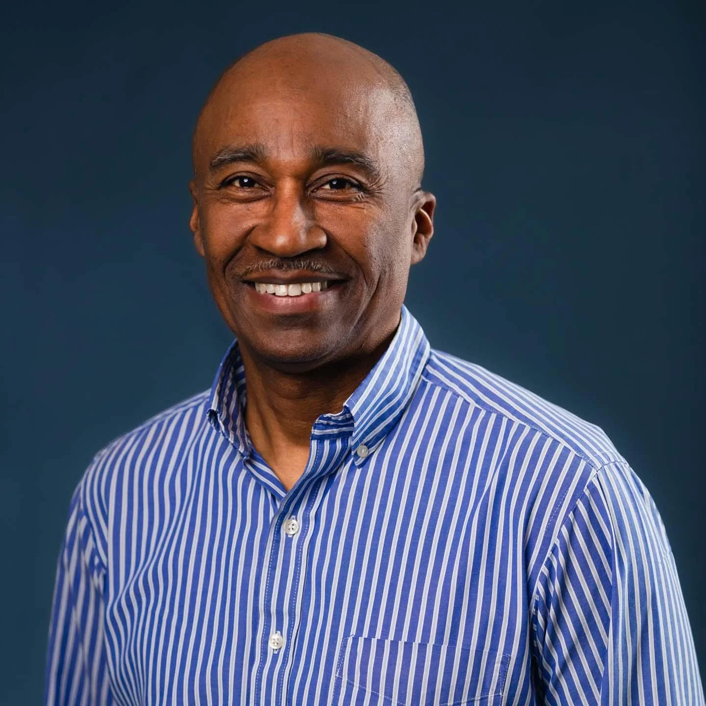 Smiling middle-aged man with a shaved head, wearing a blue and white striped button-up shirt, standing against a solid dark blue background.