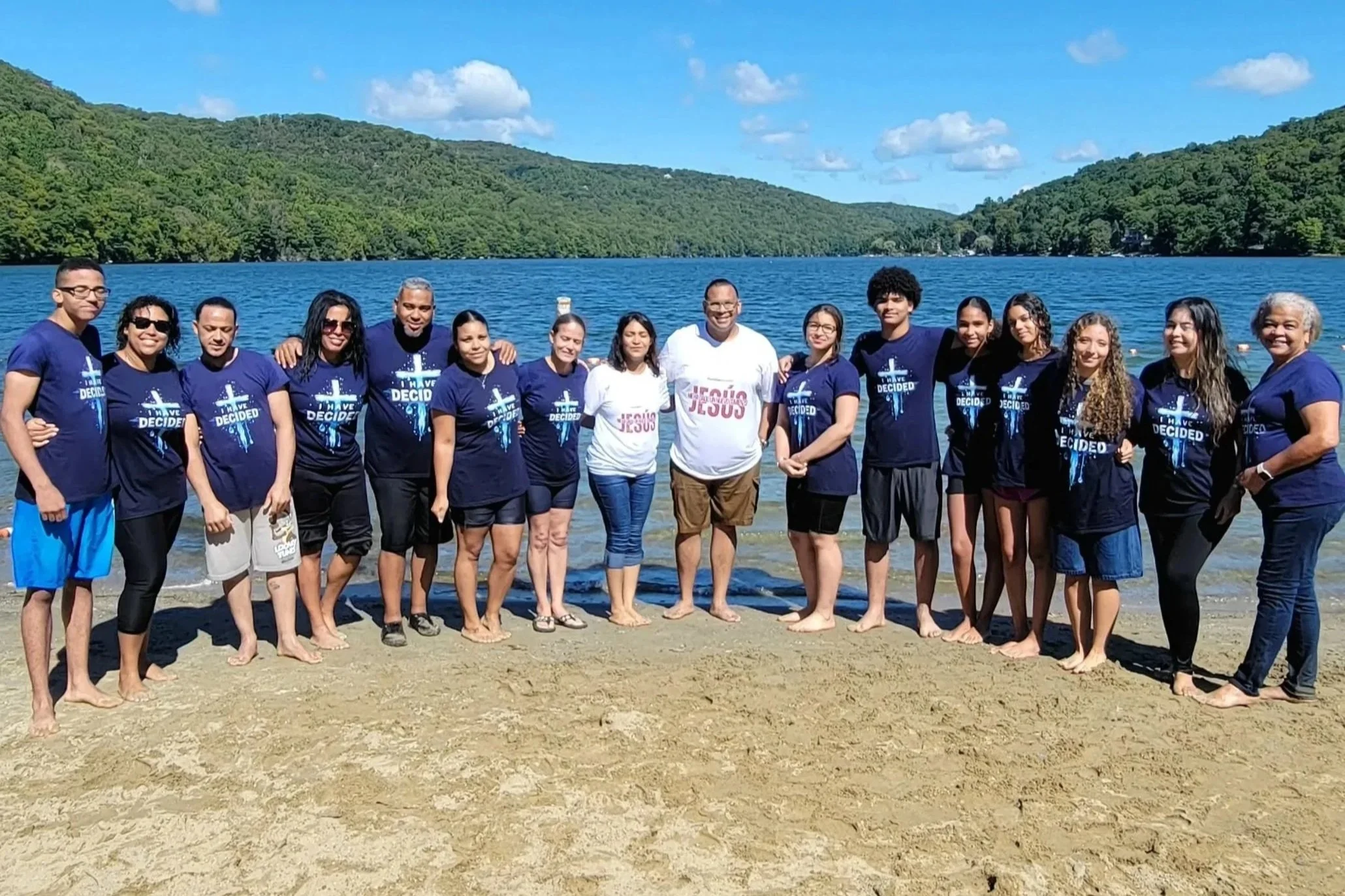 Grupo de personas en la orilla de un lago, algunas con camisetas con mensajes religiosos, rodeados de montañas verdes y cielo azul con nubes.